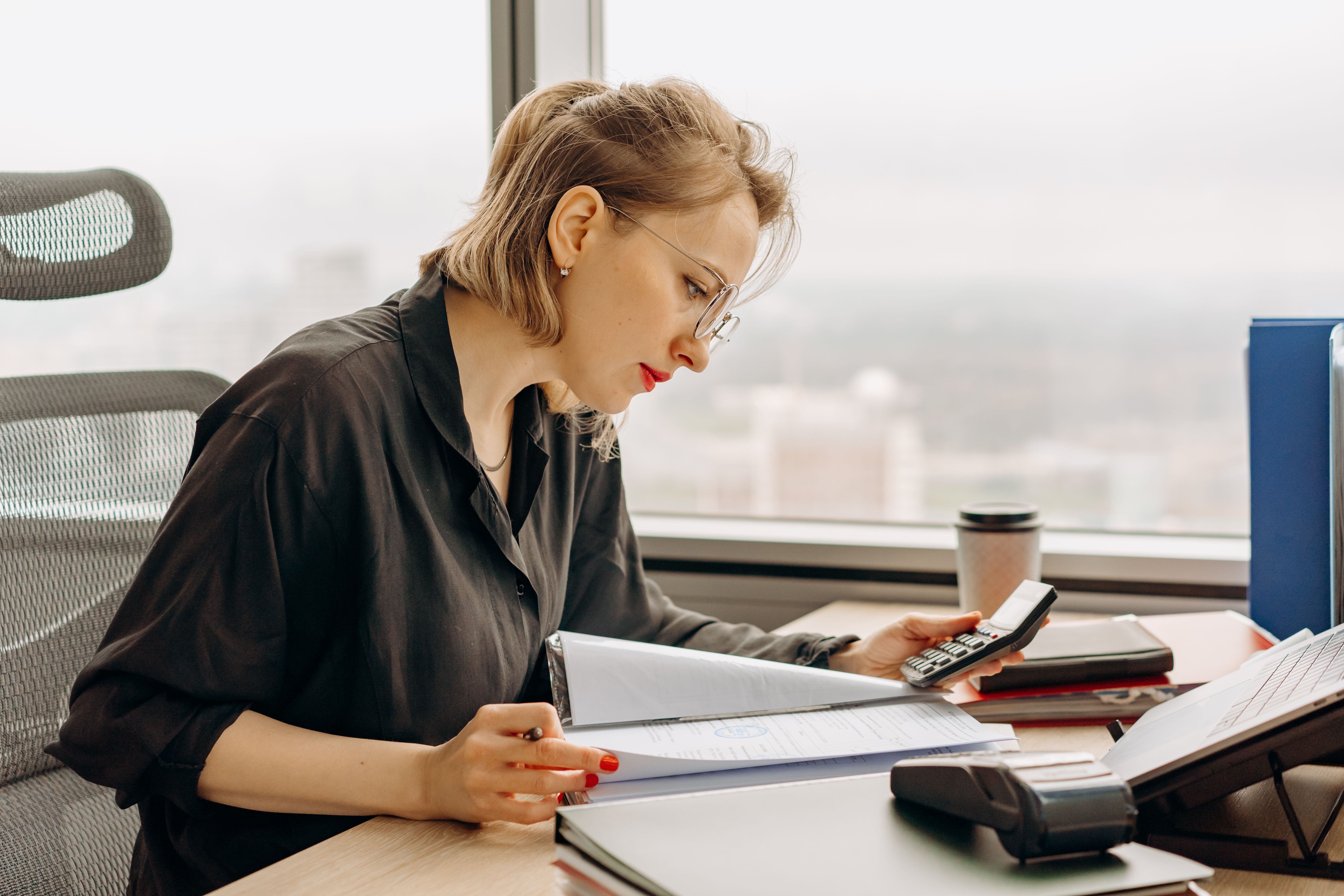 Woman in glasses working at desk using calculator and reviewing documents in office.