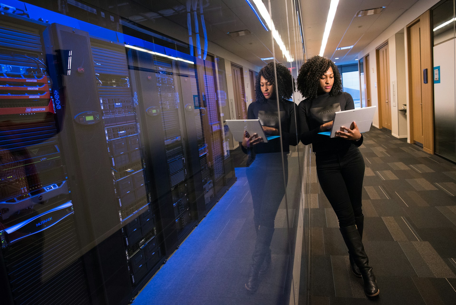 Woman in black boots using a laptop while standing by a data center server rack with blue lights.