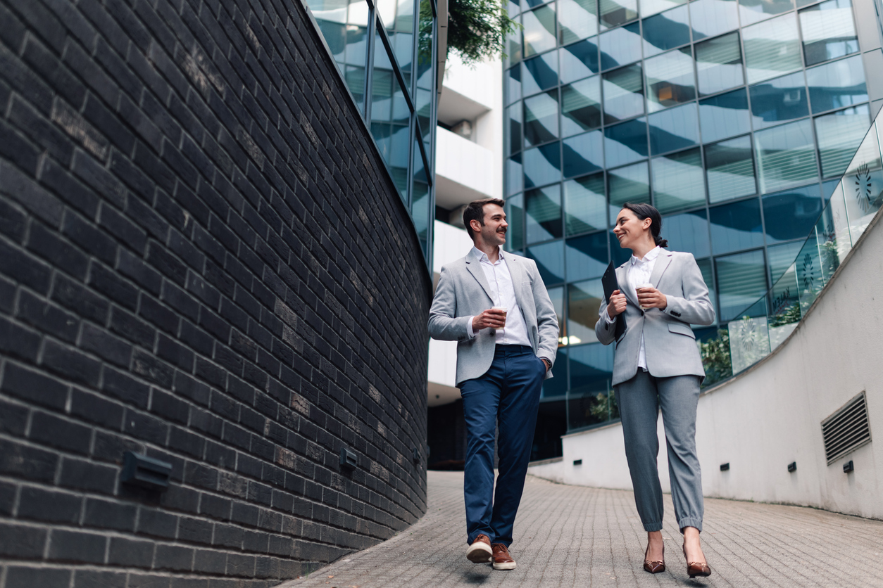 Two business professionals walking outdoors near modern office buildings holding coffee cups and a folder.