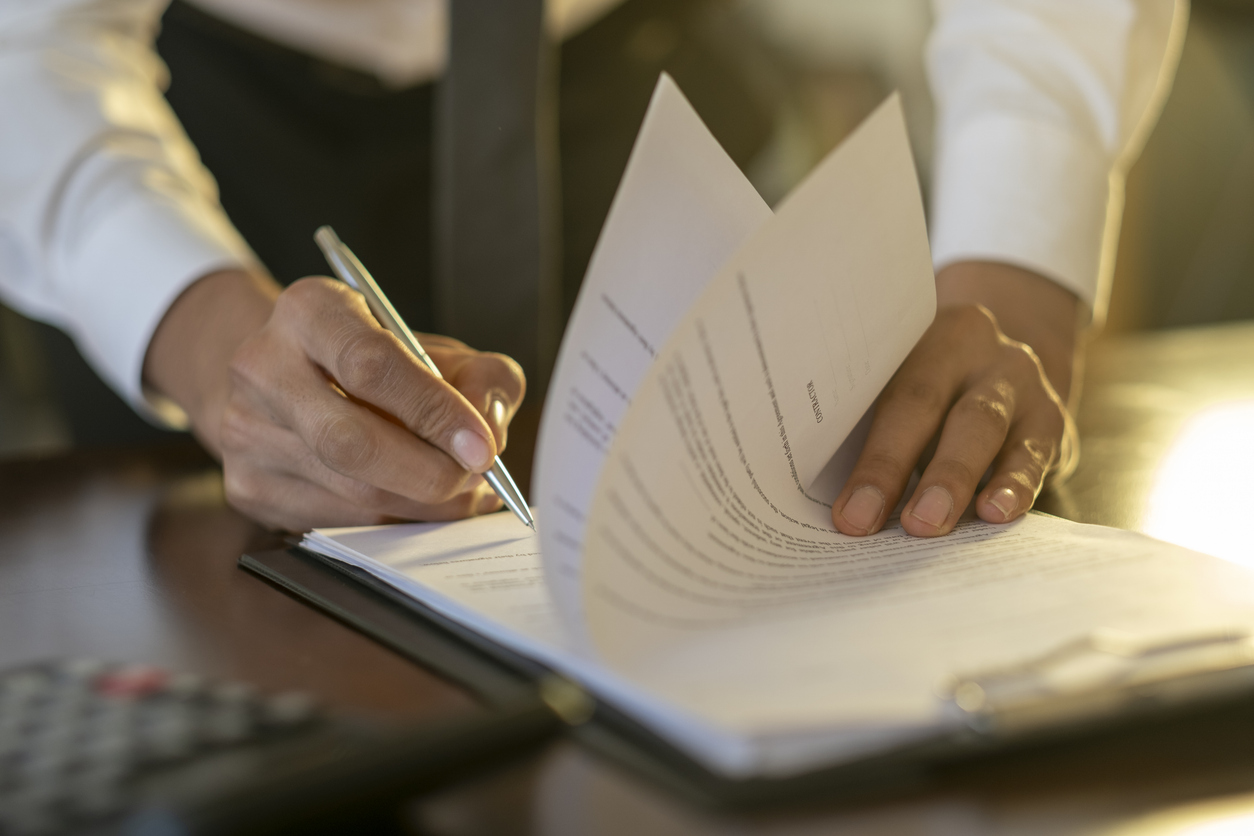Person in white shirt and tie signing or reviewing pages of a document on a desk.