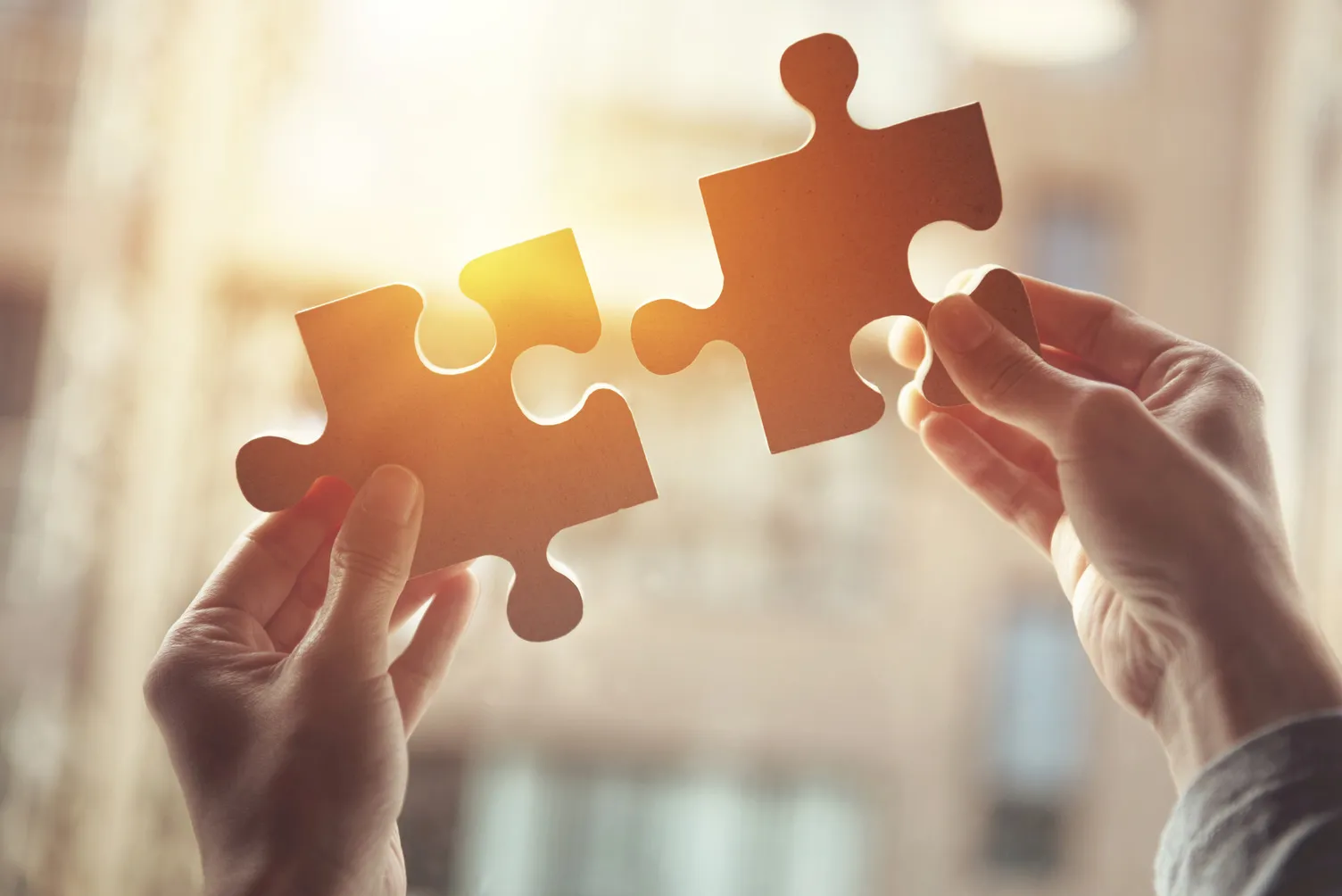 A close-up shot of two big puzzle pieces held up by two hands to a brightly lit background.