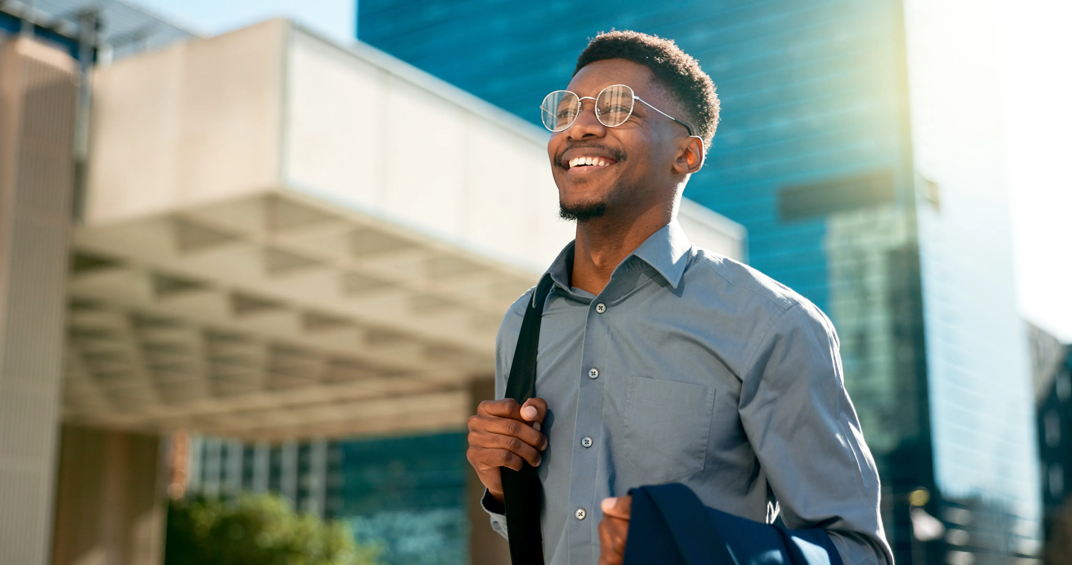 A young man with glasses  smiling while walking outside with the sun shining behind him.