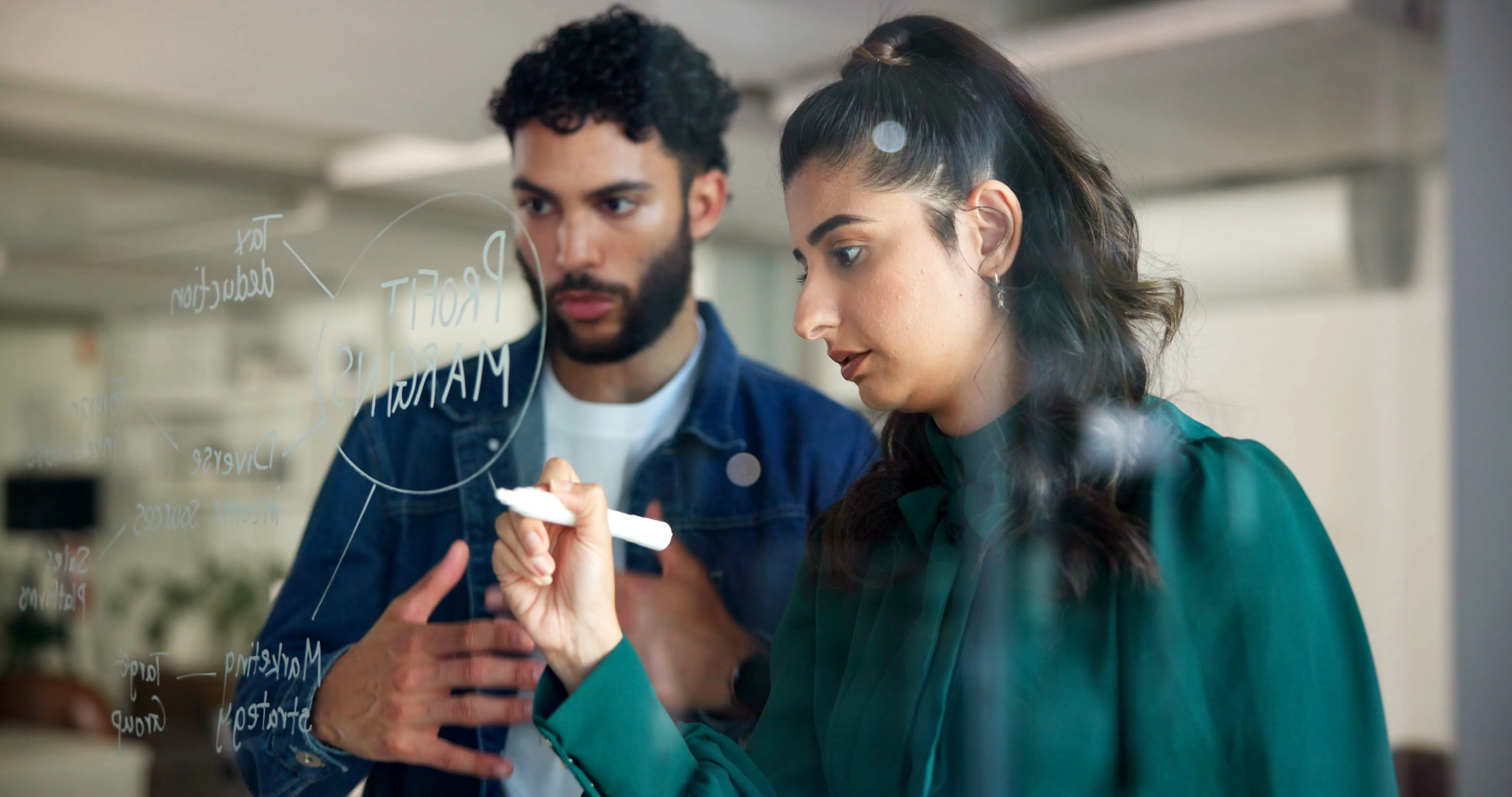 A young man and woman writing and brainstorming ideas on a clear whiteboard.