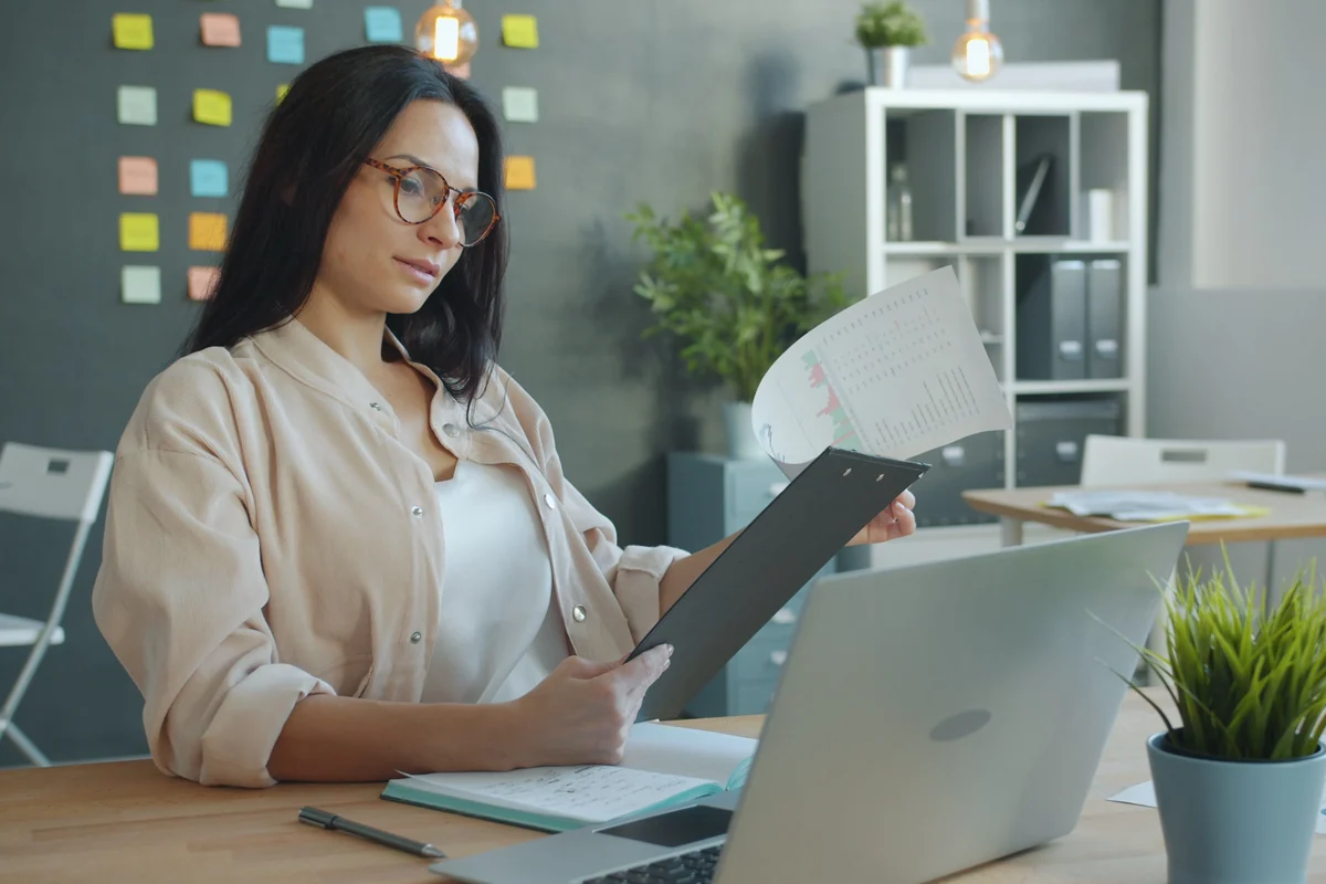 A manager reviewing a printed report at her desk beside an open laptop