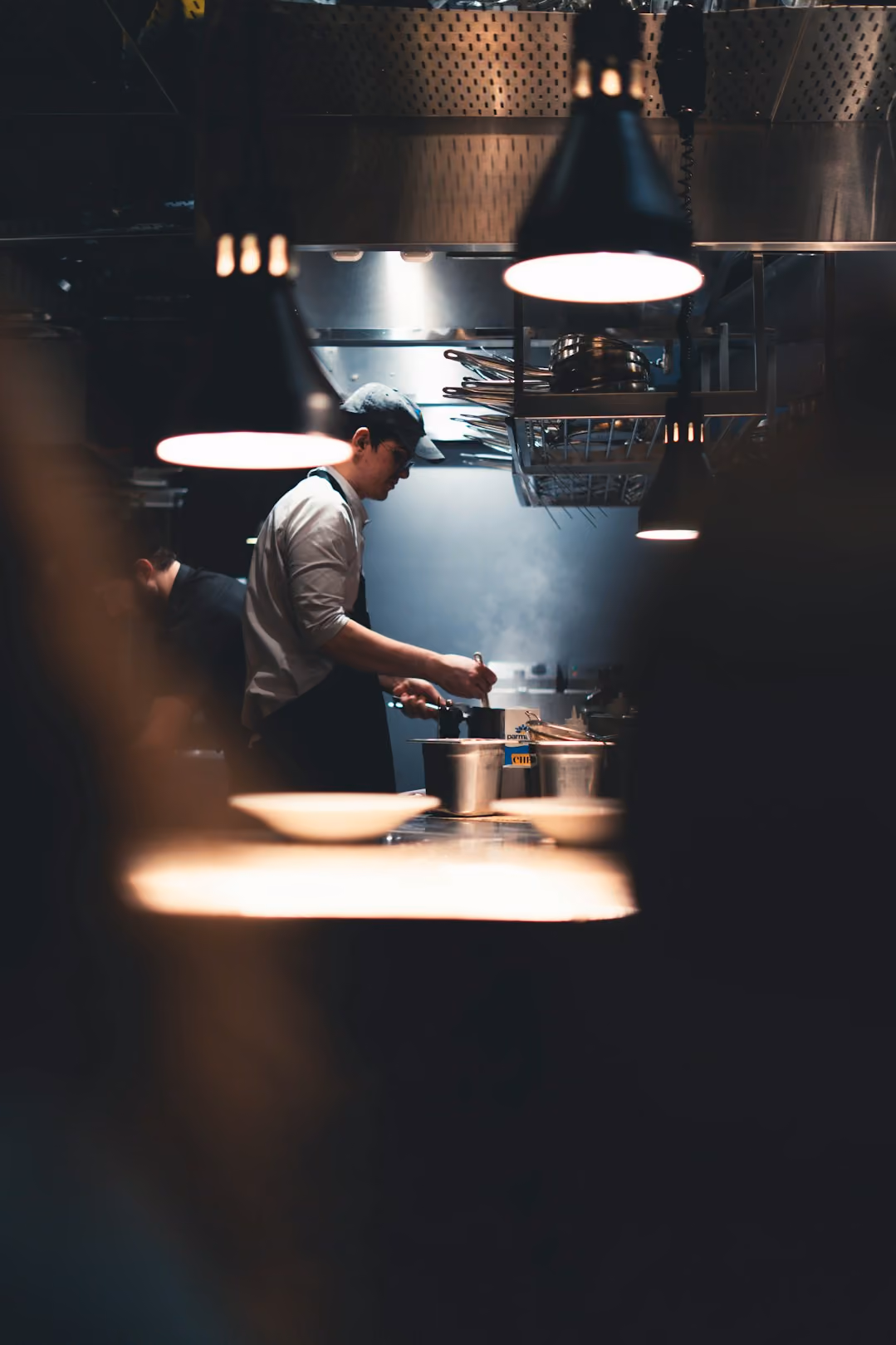 Chef wearing apron and cap, cooking in a dimly lit commercial kitchen with steam rising.