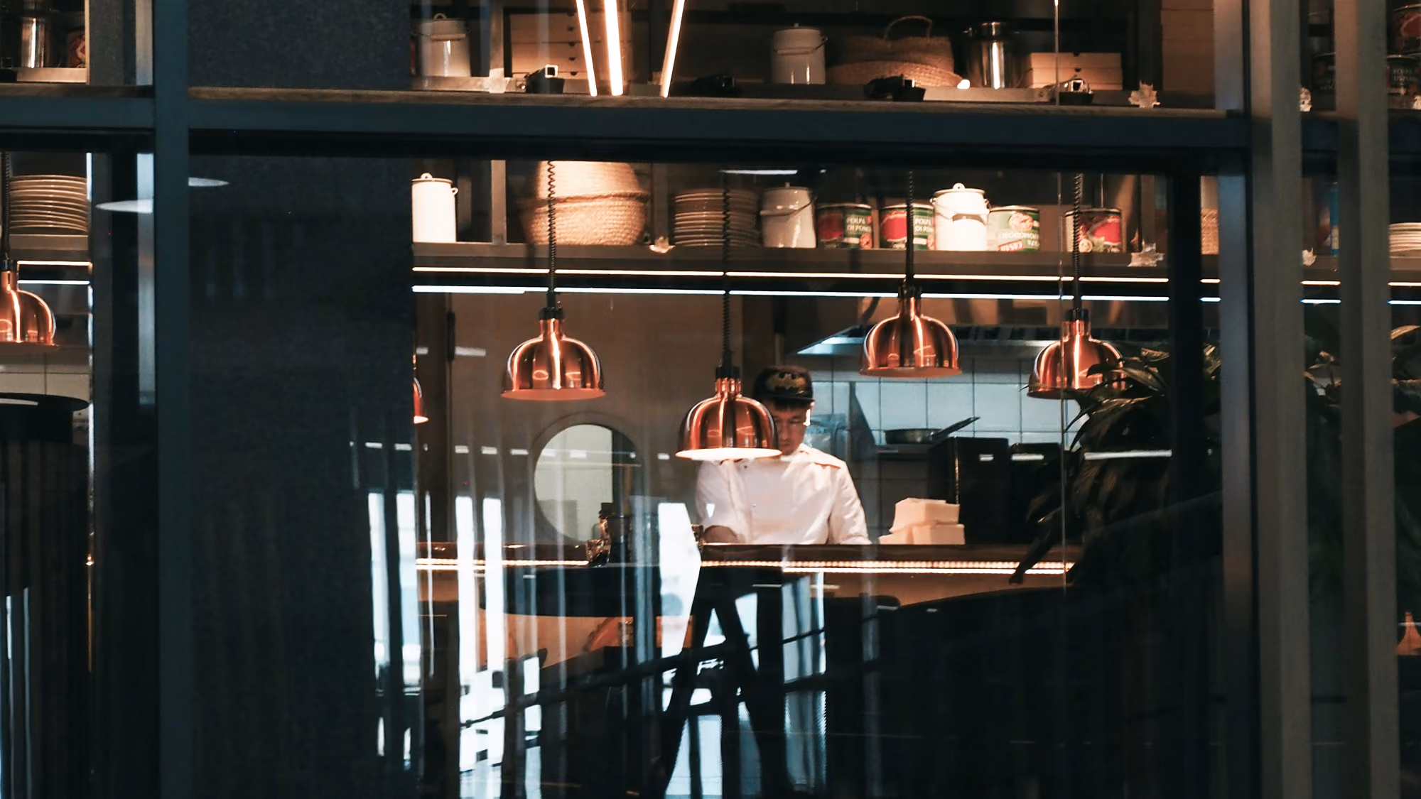 Chef in white uniform working in a modern kitchen with hanging copper lights and shelves of supplies.