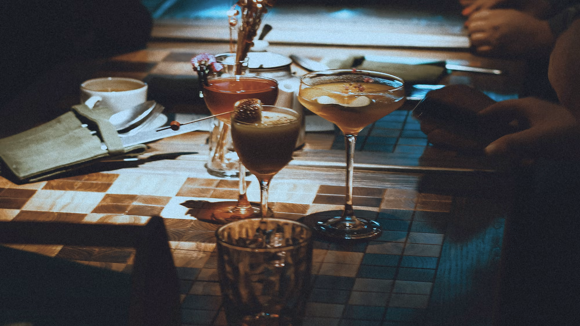 Three assorted cocktails on a patterned wooden table with hands and a cup in dim lighting.