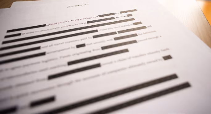 Document labeled confidential with most text heavily redacted by black bars on a wooden surface.
