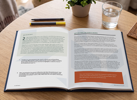 An open workbook on a table with a plant, a glass of water, and two pens