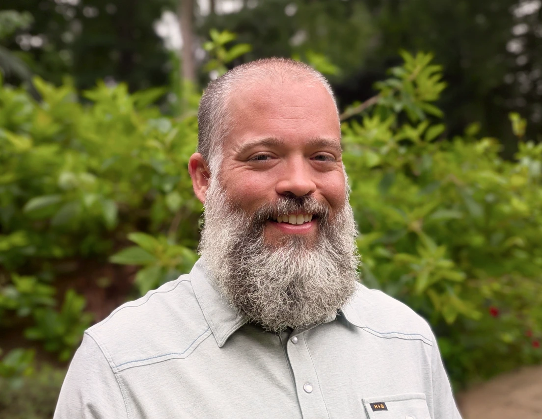 Smiling man with a full gray beard wearing a light button-up shirt outdoors with green foliage behind.