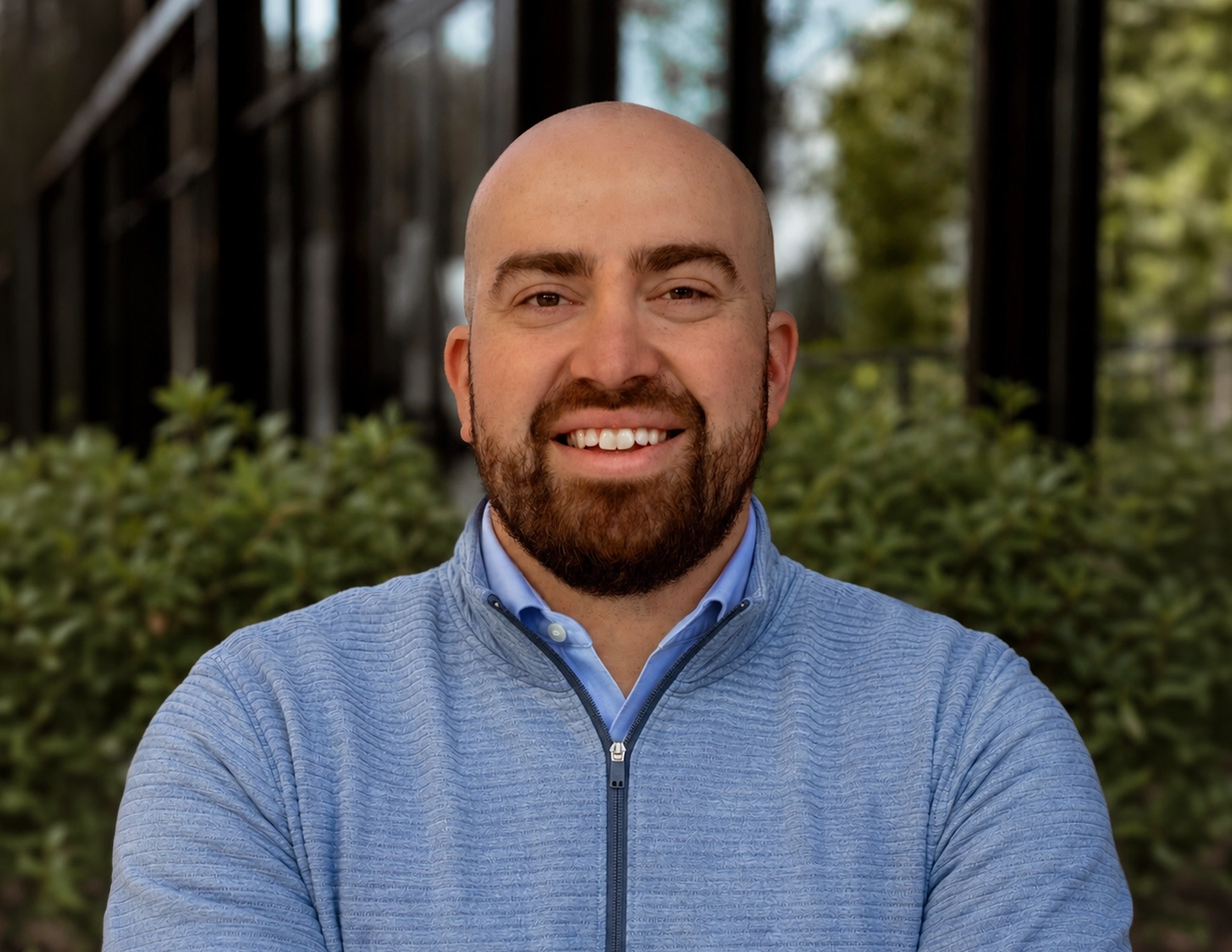 Smiling bald man with beard wearing a blue zip-up sweater outdoors with greenery in the background.