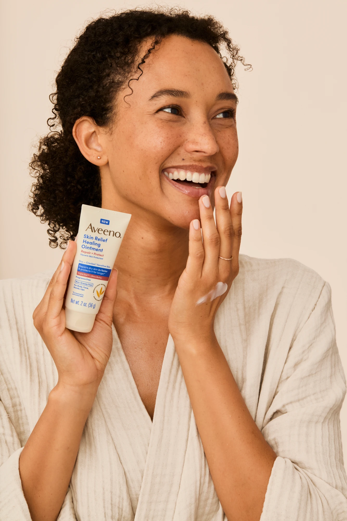 A smiling model holding an Aveeno tube while touching her cheek.