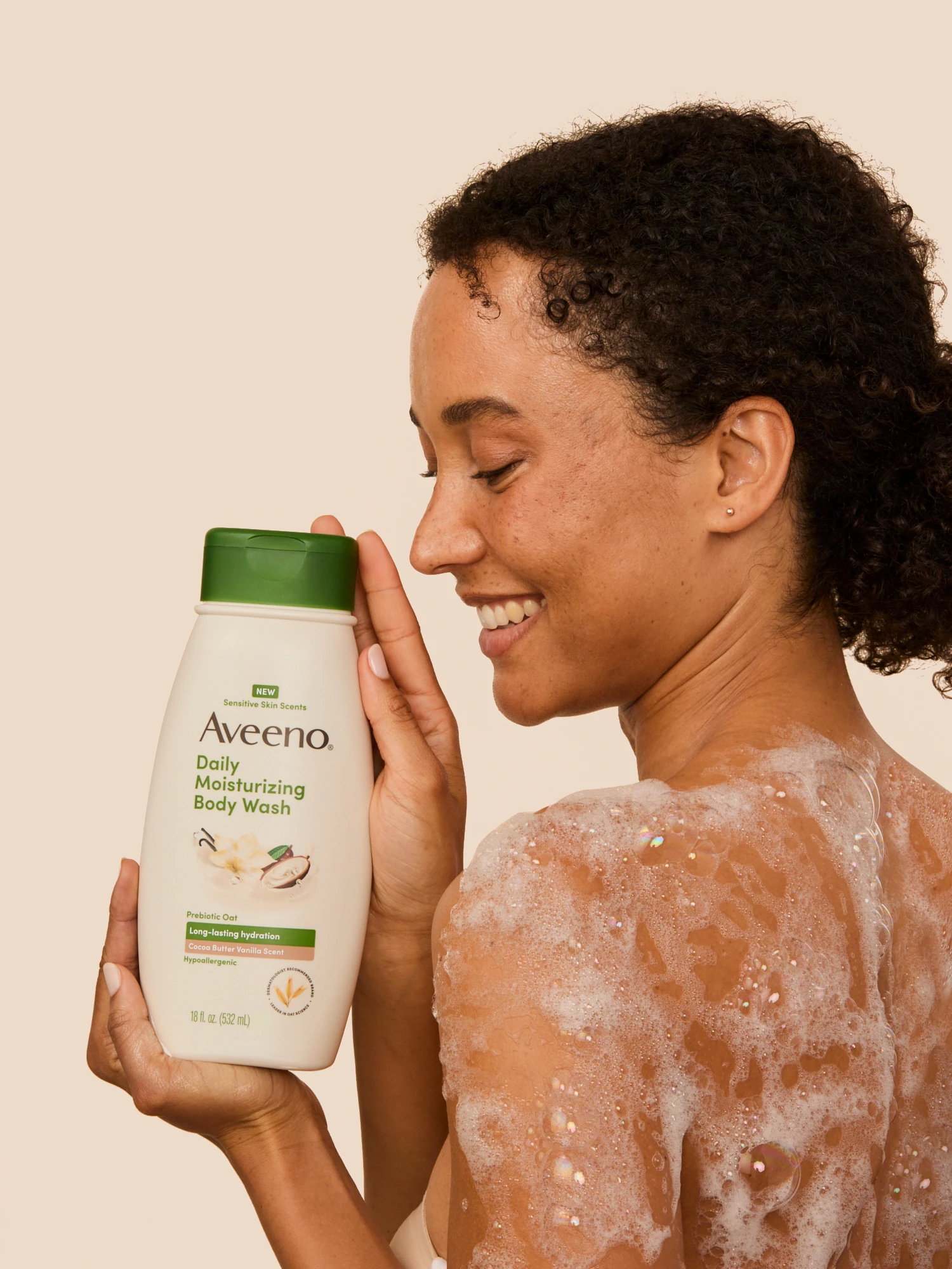 A smiling model holding an Aveeno body-wash bottle beside her lathered shoulder.