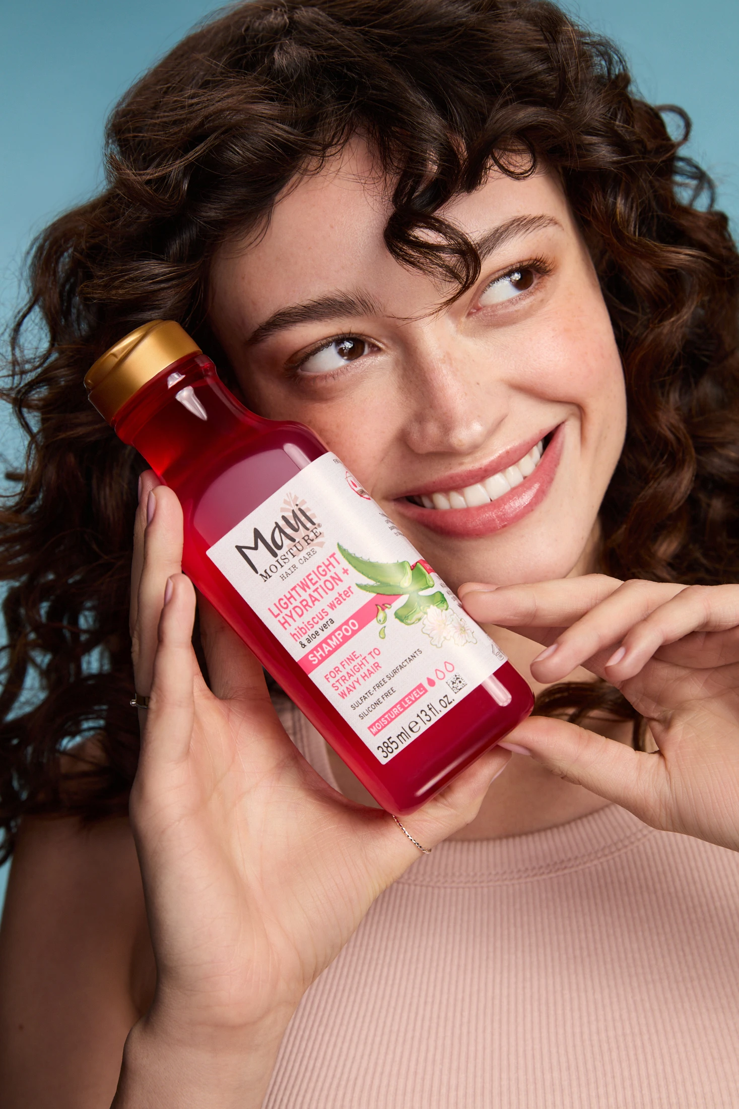 A smiling model holding a pink Maui Moisture bottle beside her cheek.