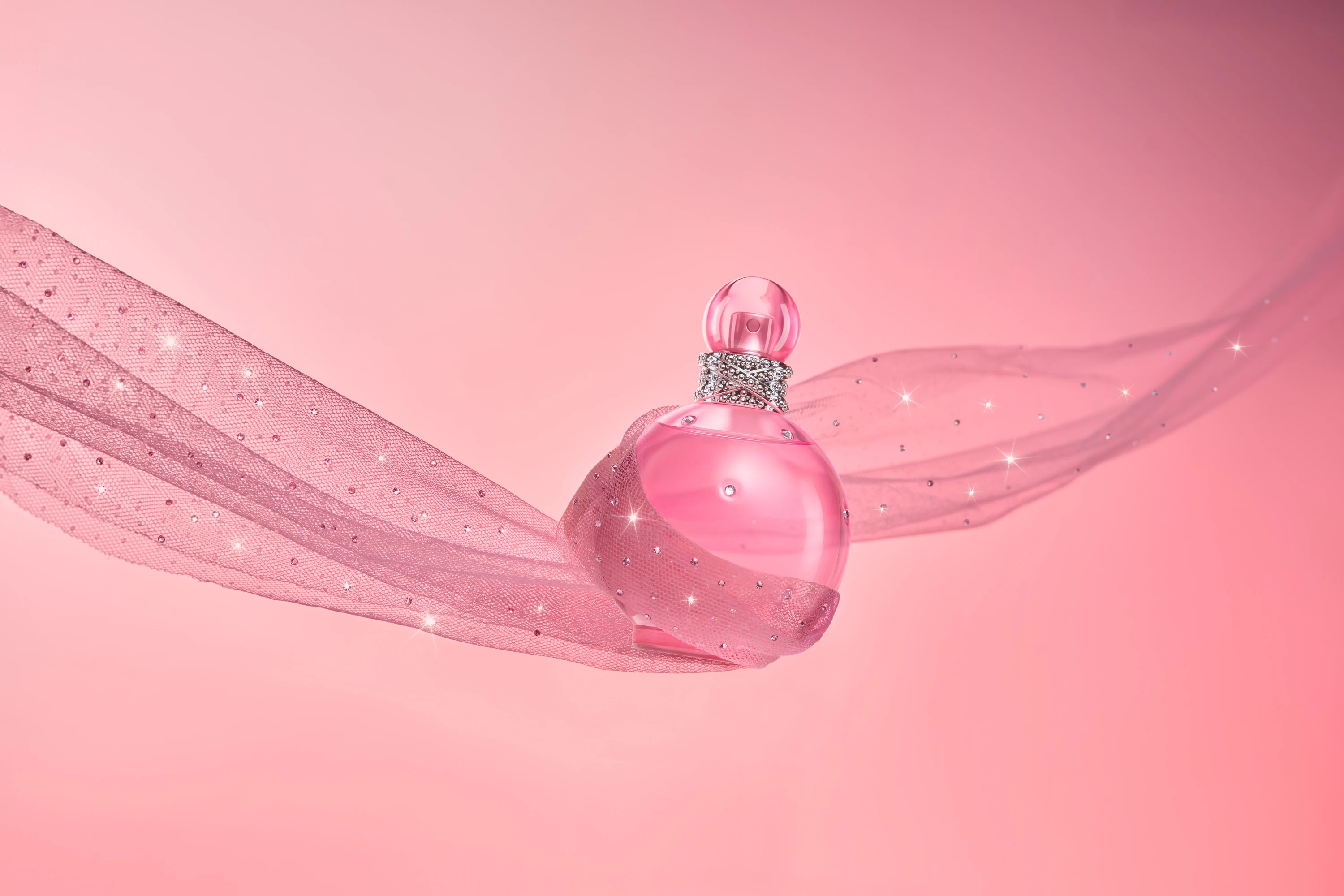 A pink round fragrance bottle suspended over flowing pink fabric.