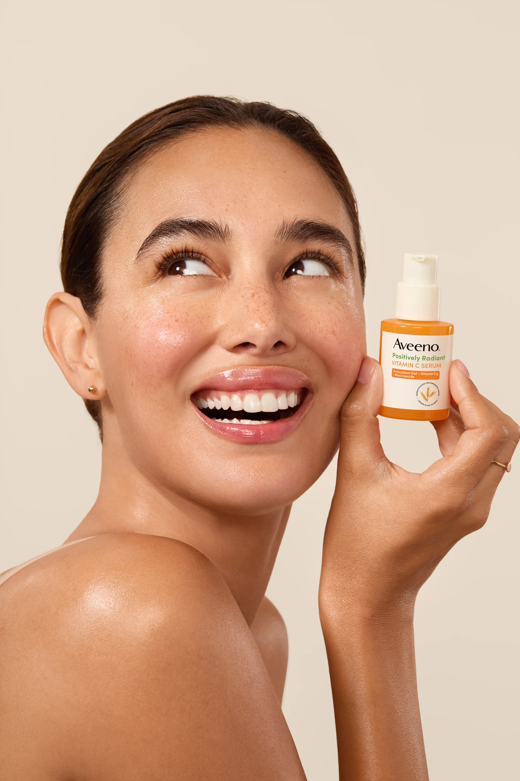 A smiling model holding an Aveeno serum bottle beside her face.