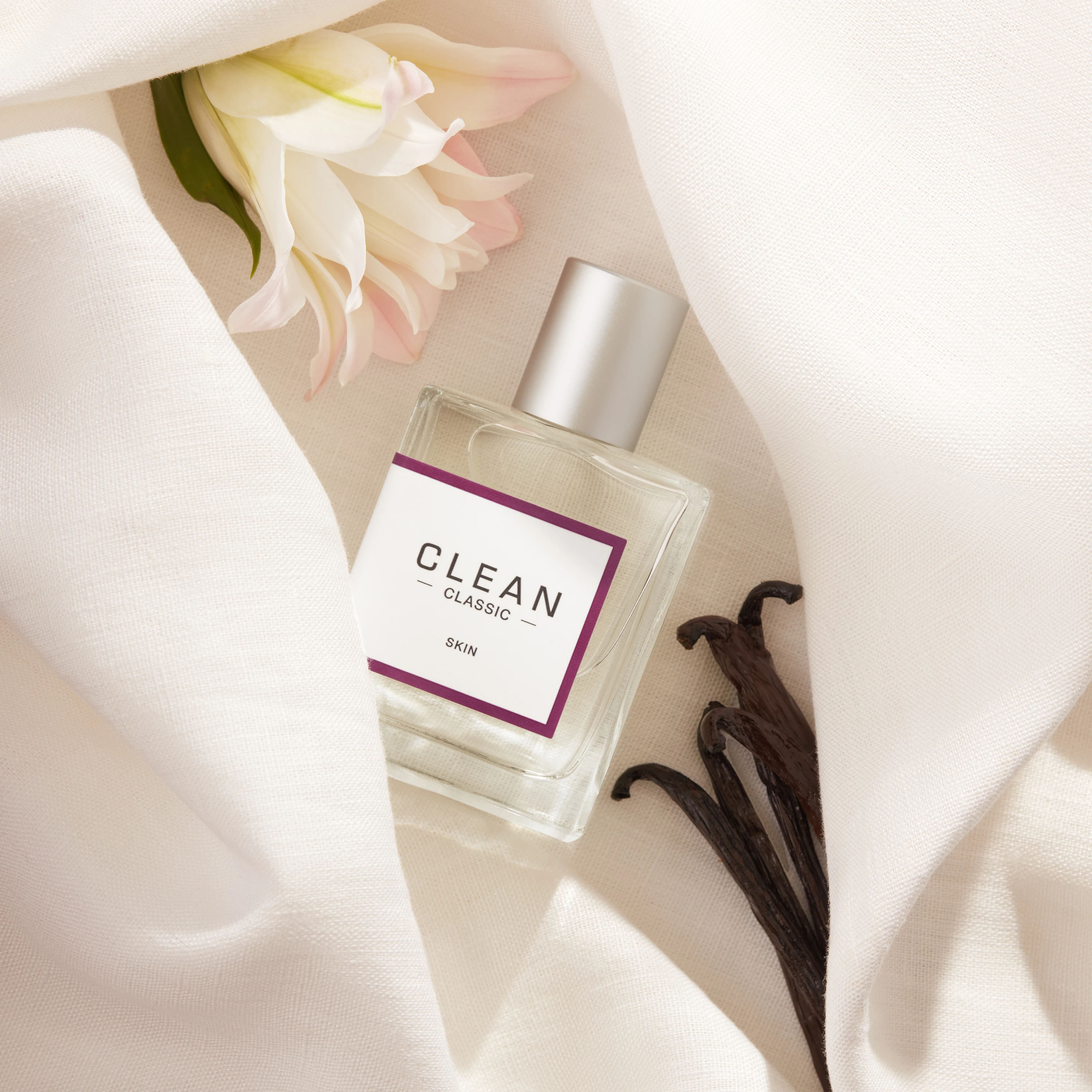 A Clean fragrance bottle on pale fabric beside flowers and vanilla pods.