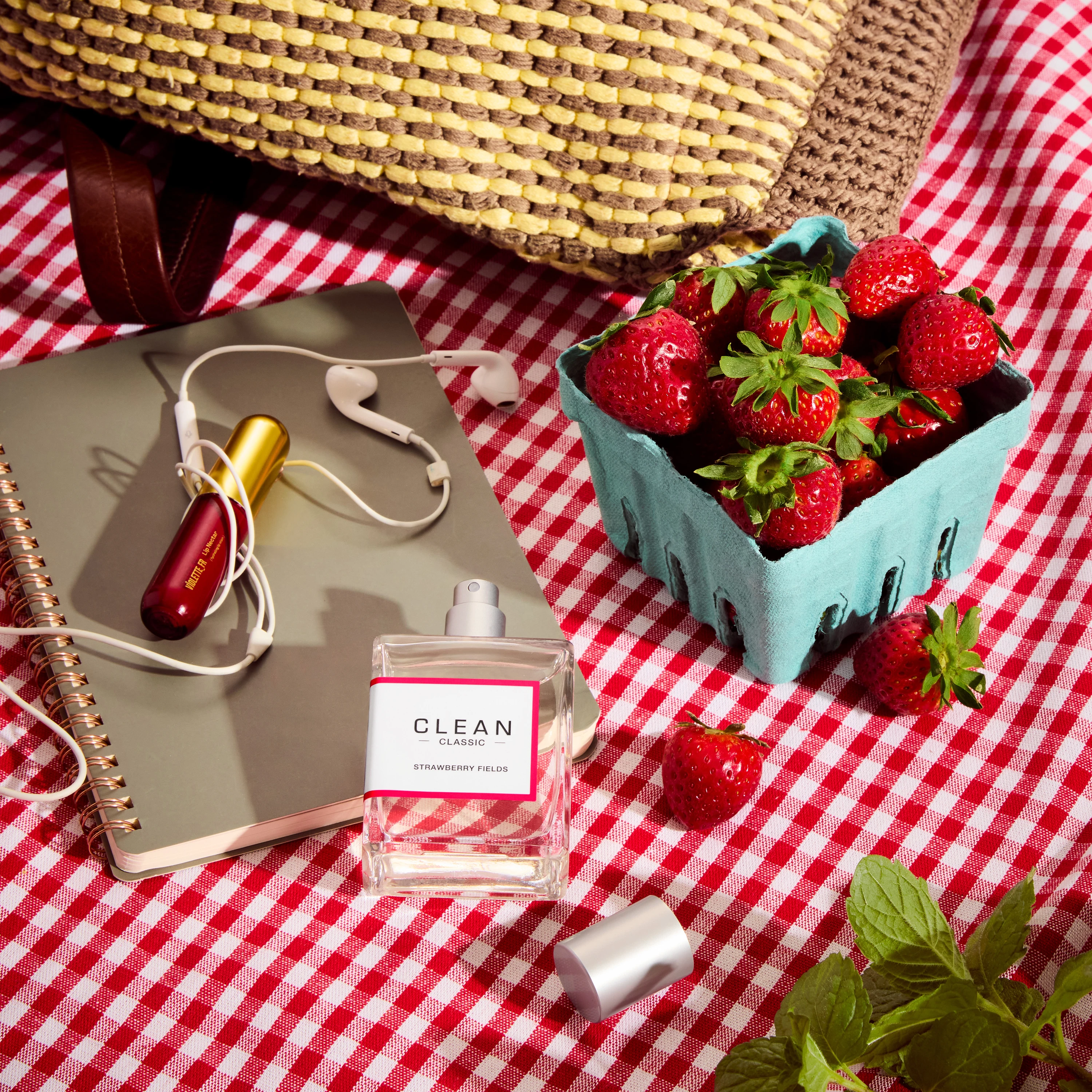 A pink Clean fragrance bottle beside strawberries on a picnic blanket.