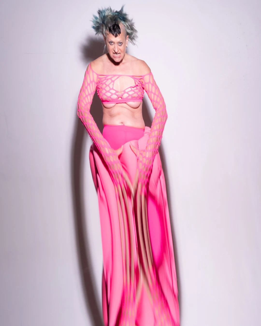 Peaches posing in a long pink outfit against a pale wall.