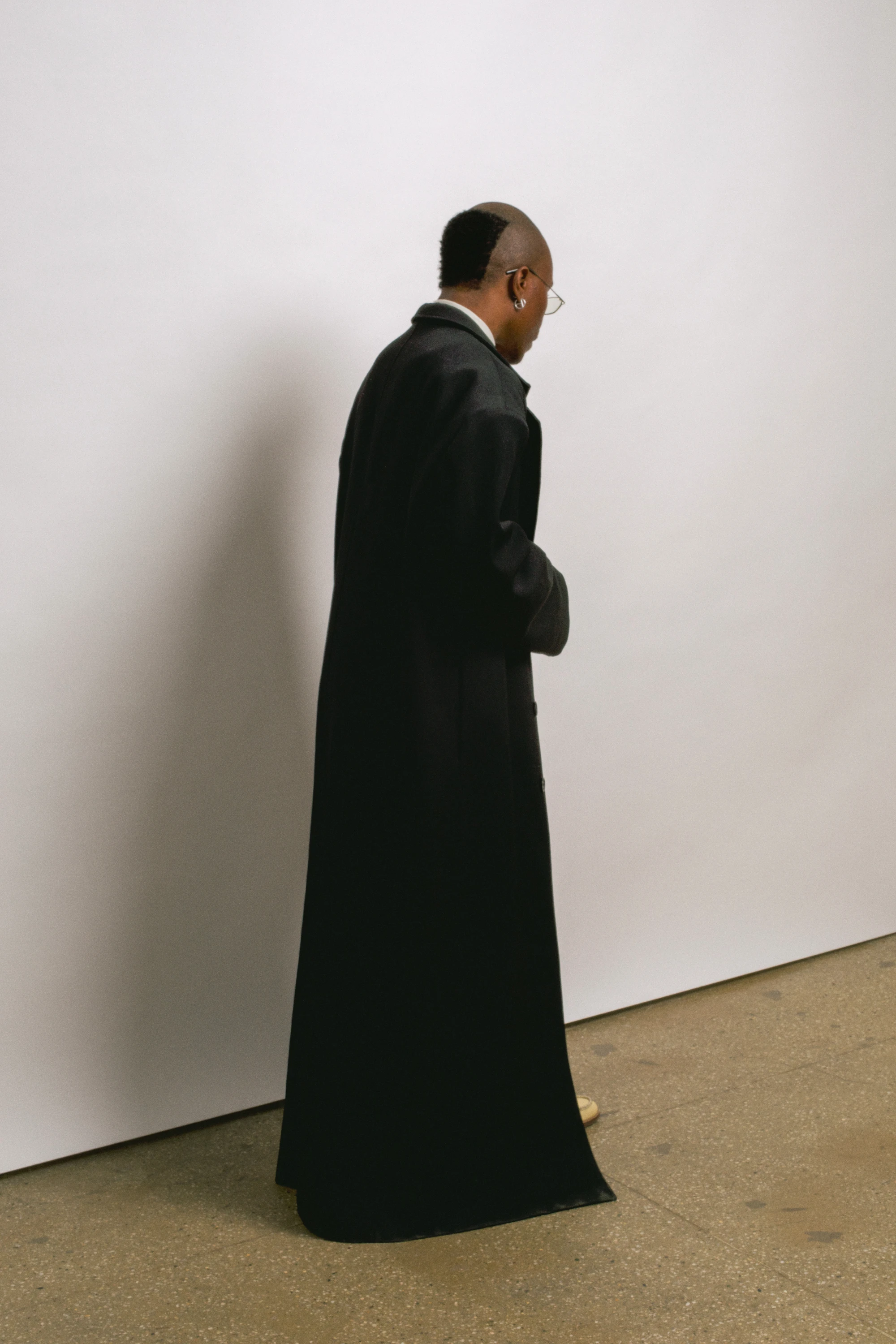 TYGAPAW standing in a long black coat beside a pale wall.