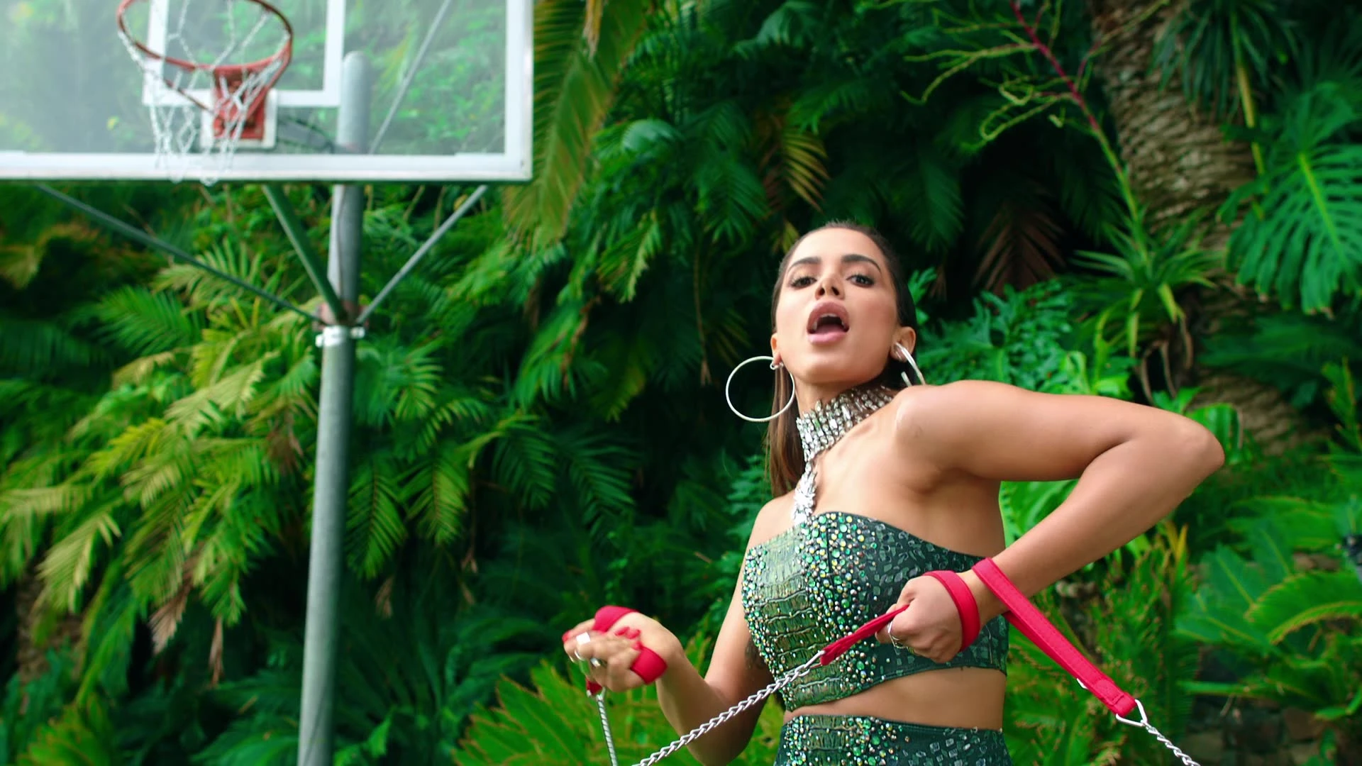 Anitta posing beneath a basketball hoop among tropical plants.