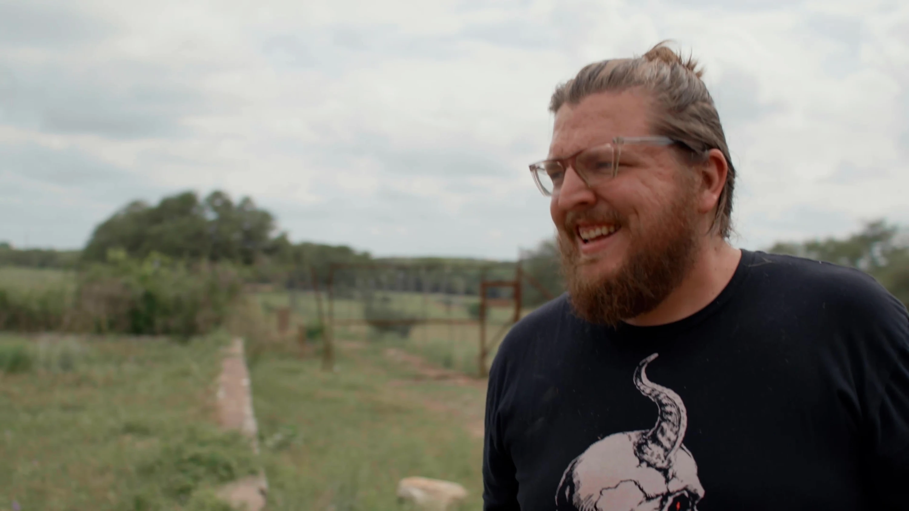 A bearded person standing in a grassy field.