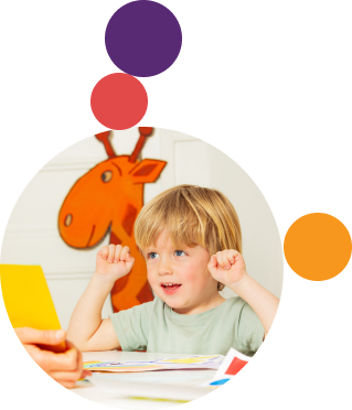 Young boy with blonde hair raising fists while sitting at a table, orange giraffe drawing in background.