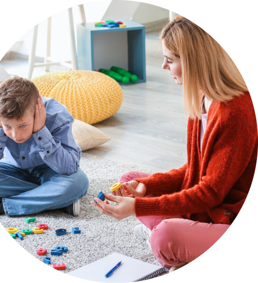 Woman speaking to upset boy covering his ears while sitting on a rug with scattered toys.