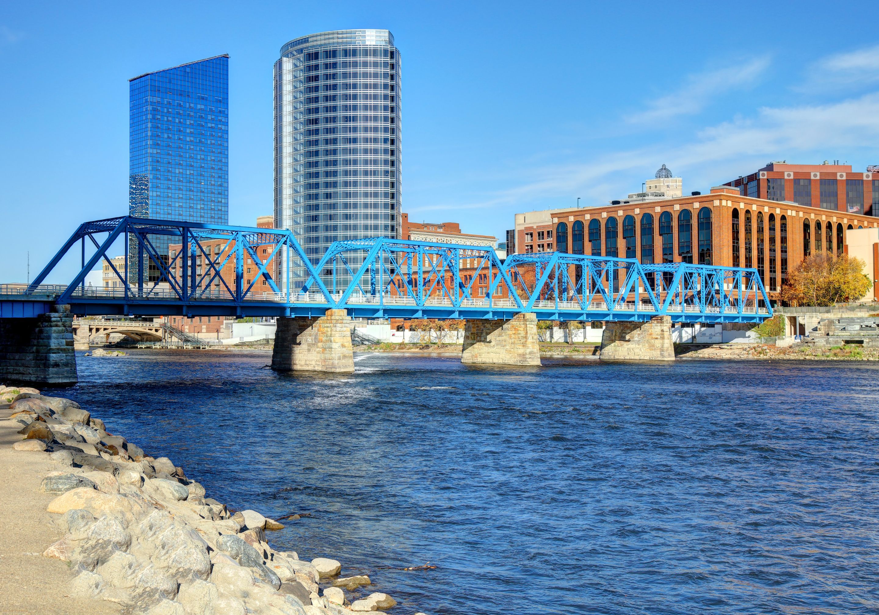 Blue truss bridge over a wide river with rocky shoreline, modern glass and brick buildings in the background under a clear blue sky.