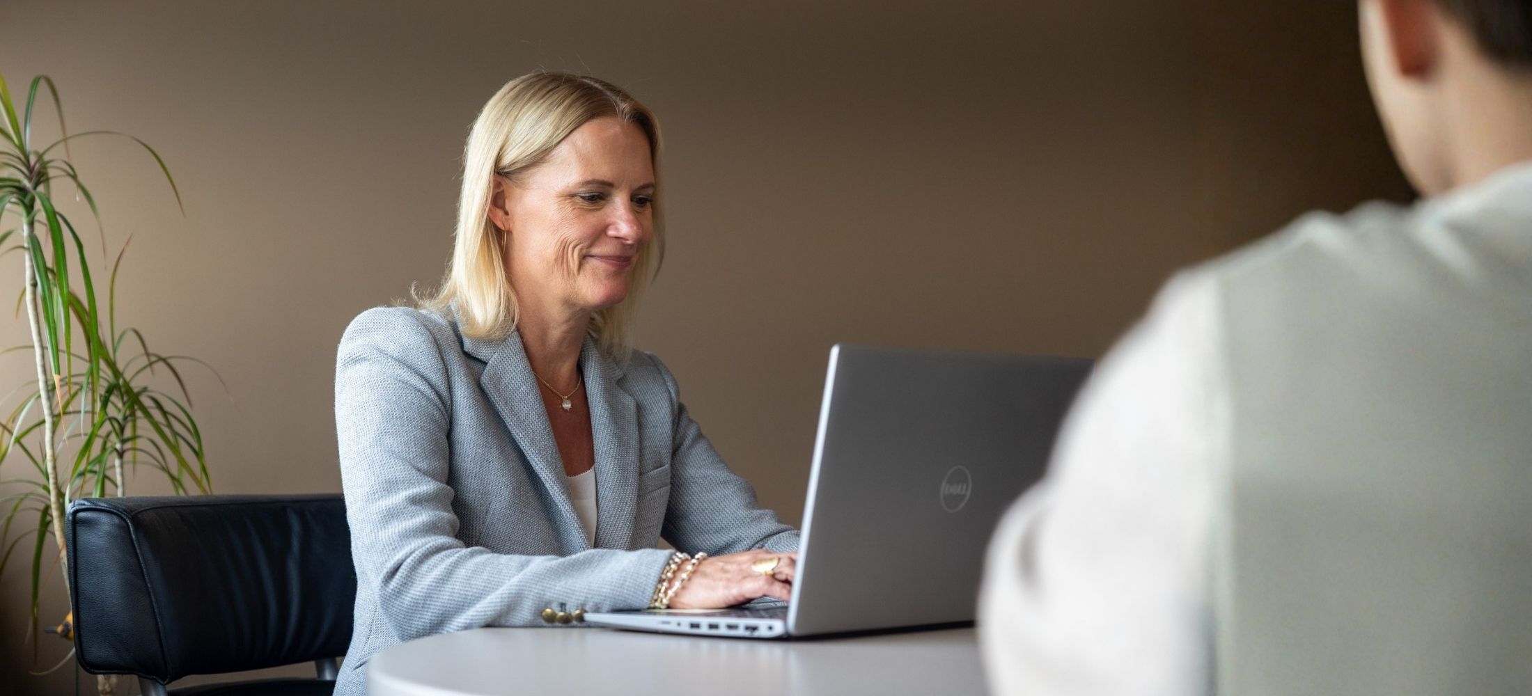 Smiling blonde woman in a grey blazer working on a silver Dell laptop at a round table with a person partially visible in front, against a plain brown background with a tall green plant on the left.