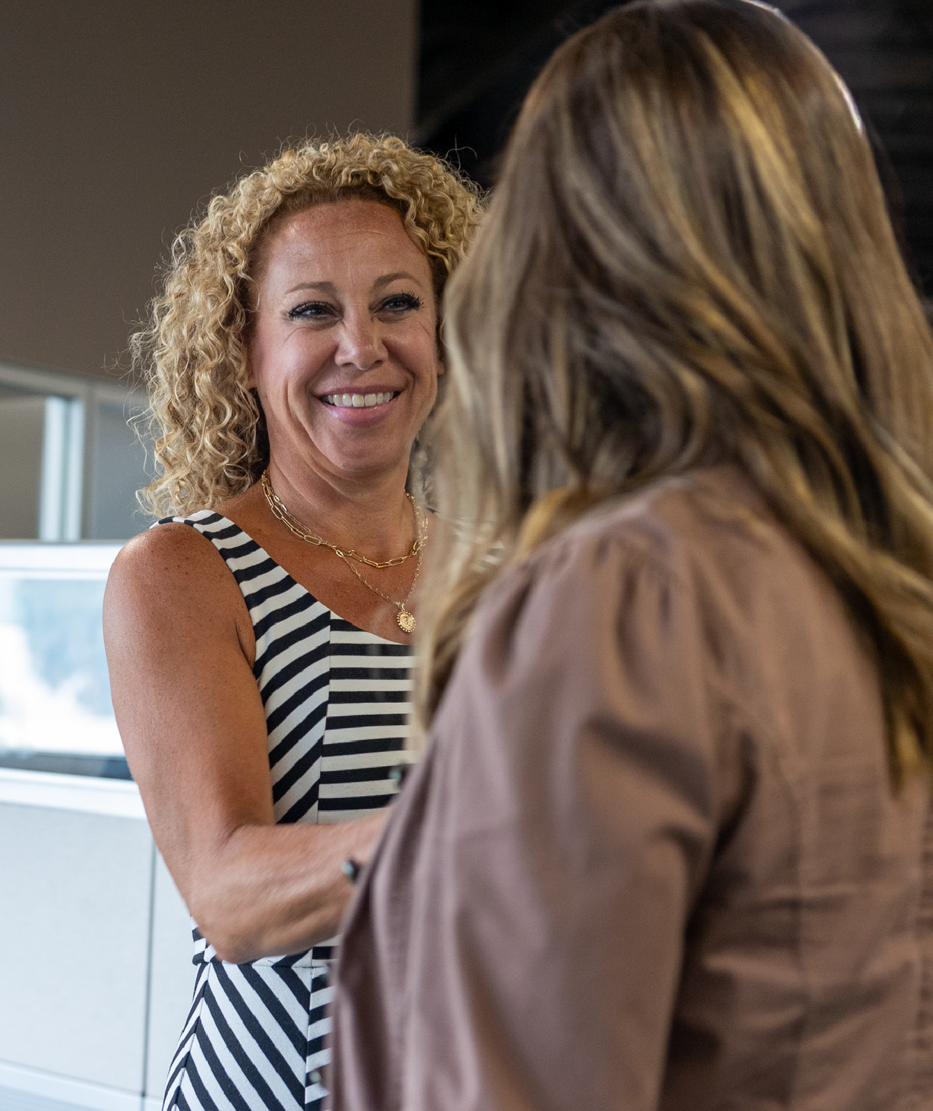 Smiling woman with curly blonde hair wearing a black and white striped sleeveless dress shaking hands with another woman with long light brown hair.