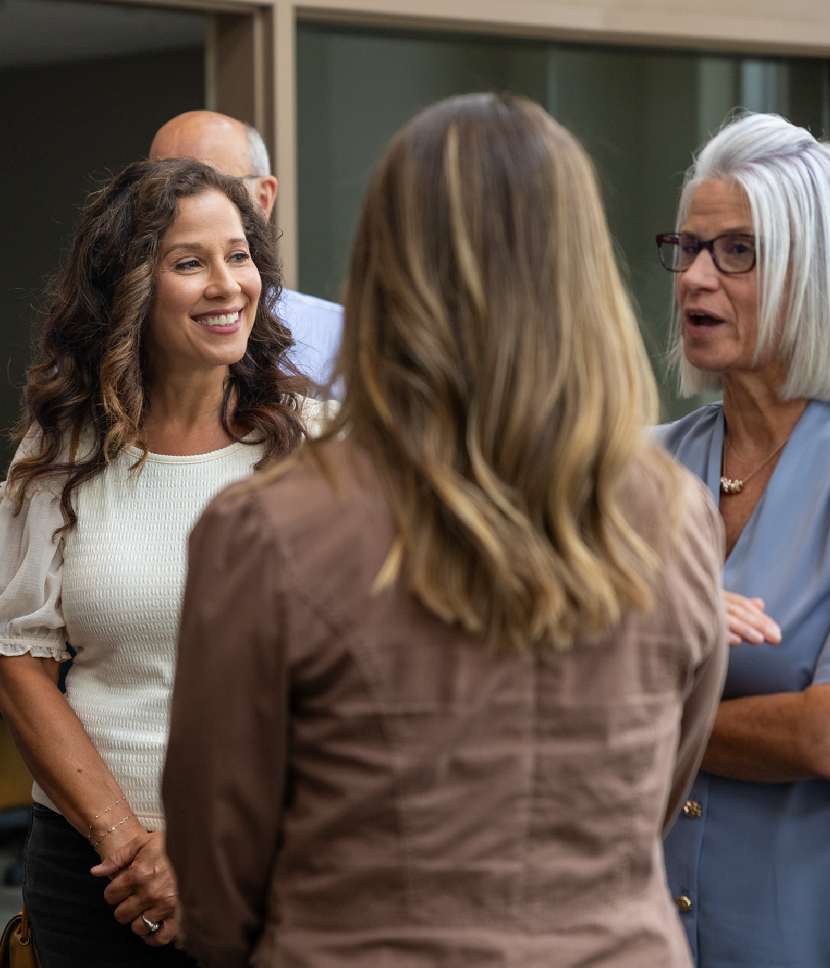 Four adults engaged in a conversation in a casual indoor setting; a smiling woman with curly hair in a white top, a woman with shoulder-length blonde hair wearing a brown jacket seen from behind, a woman with short white hair and glasses in a blue blouse speaking, and a man with balding hair partially visible in the background.