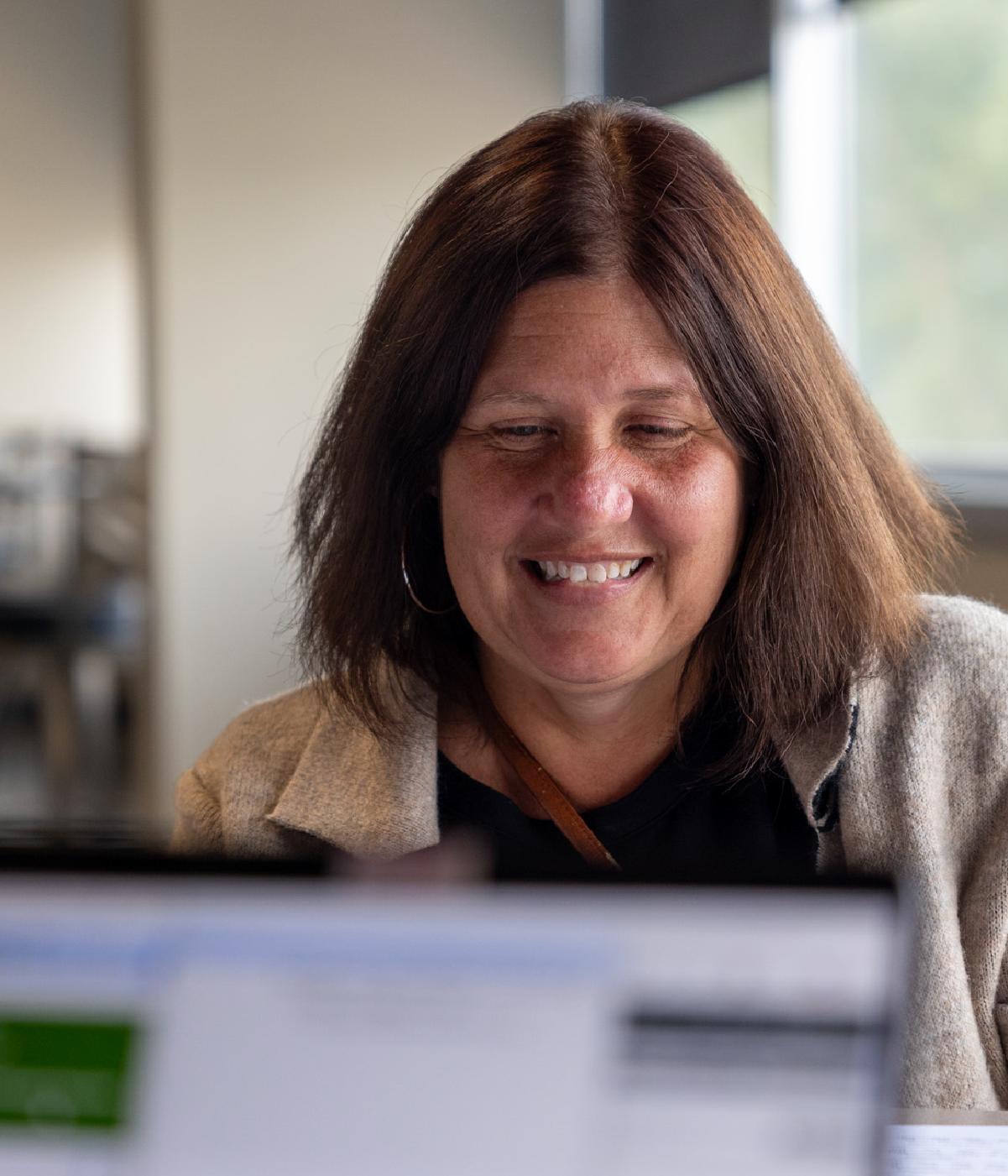 Smiling woman with shoulder-length brown hair sitting indoors, looking at a computer screen with blurred content in the foreground.
