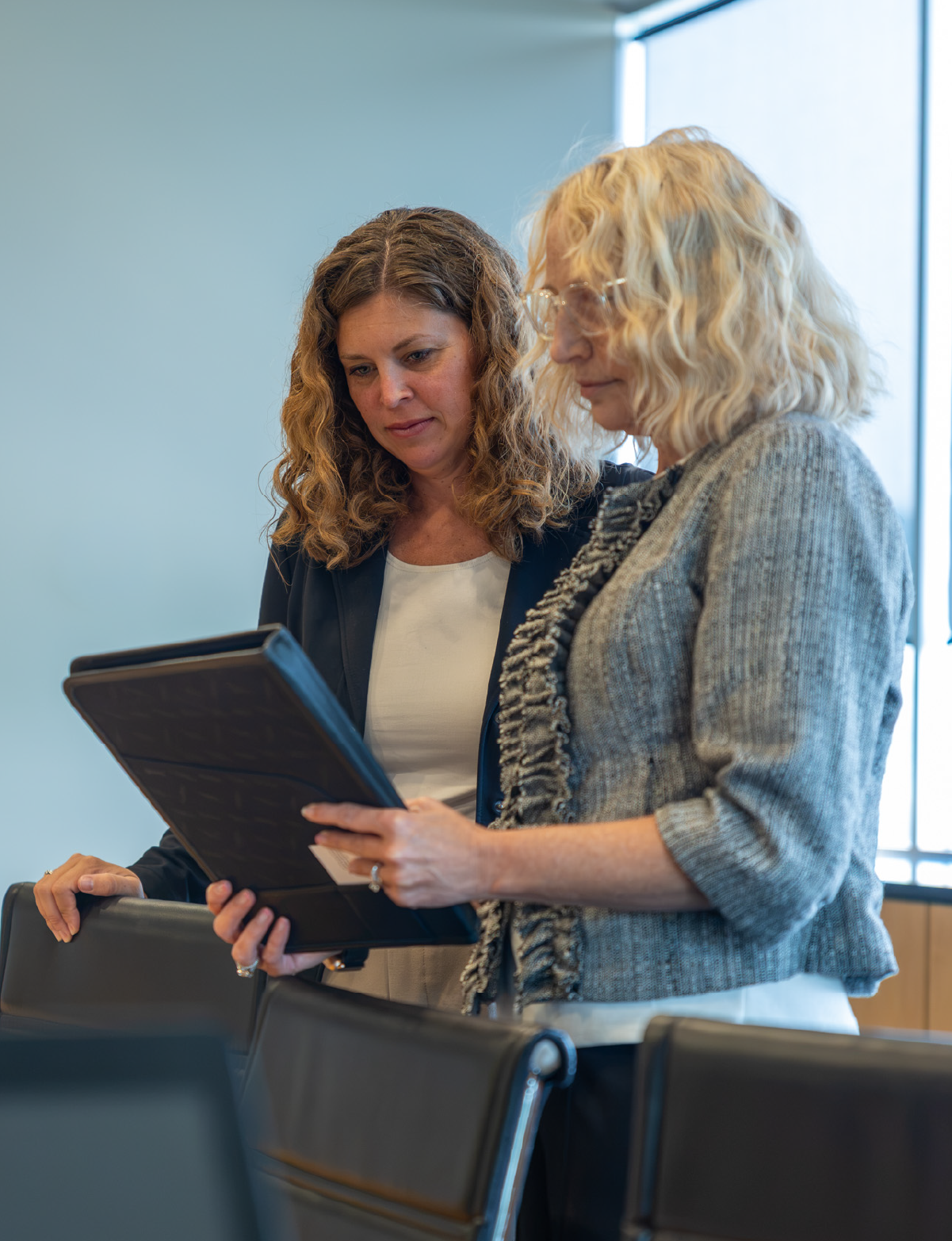 Two professional women standing in an office conference room, looking at a tablet together, with rows of black chairs in the foreground.