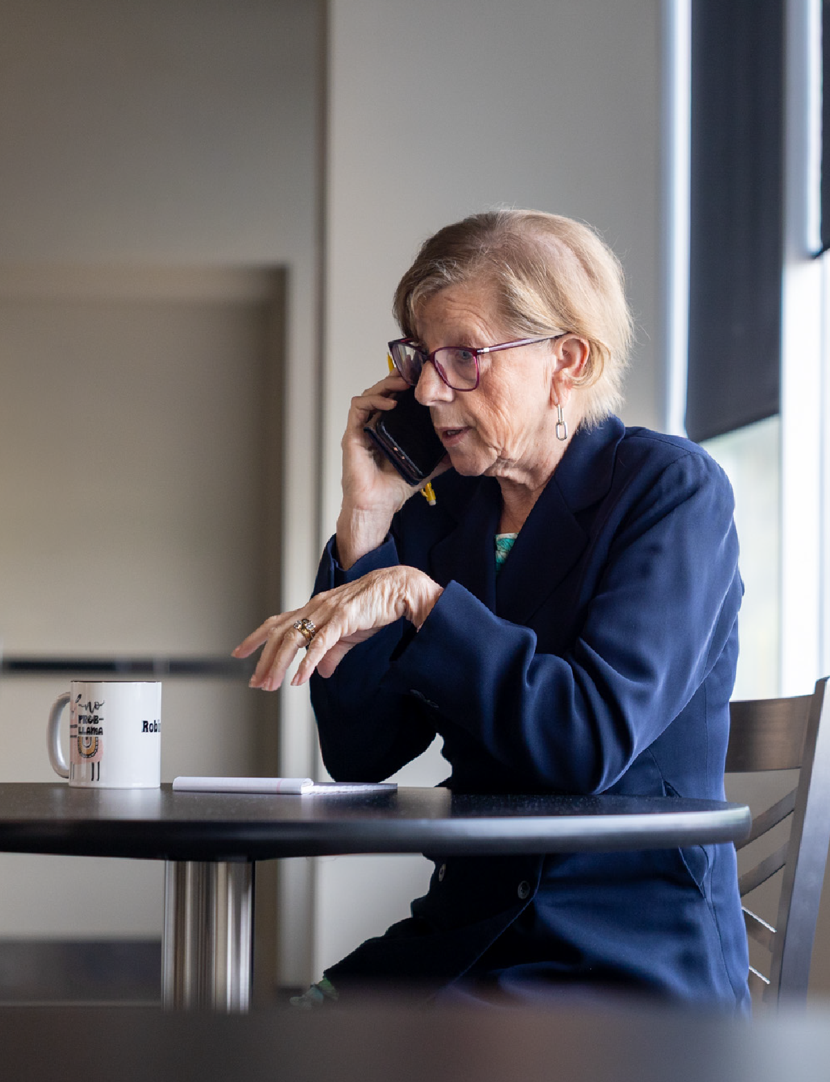 Older woman with short light hair and glasses wearing a navy blue blazer is sitting at a round table, talking on a phone held to her ear, with a white mug and notebook on the table.