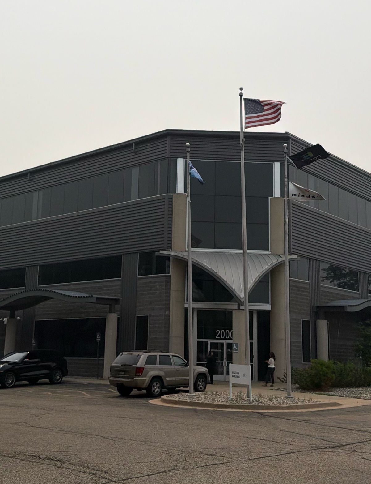 Modern two-story commercial building with large dark glass windows, a metal canopy over the entrance, and an address number 2000 above the door, three flagpoles with an American flag and two other flags, two vehicles parked near the entrance, and two people standing outside.
