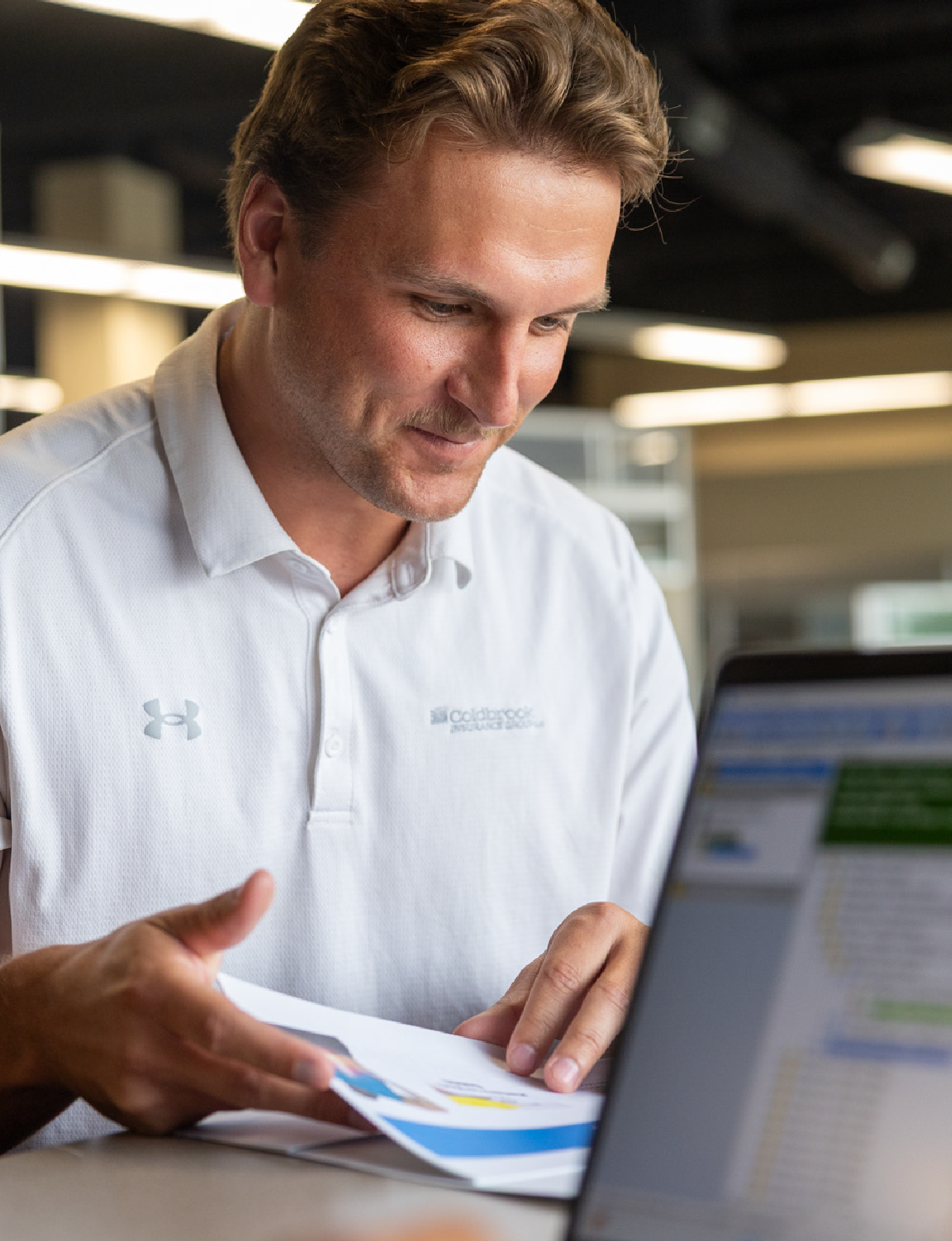Man with short brown hair wearing a white Coldbrook Insurance Group polo shirt, looking down and smiling while holding printed documents, with a blurred laptop screen in the foreground in an office setting.