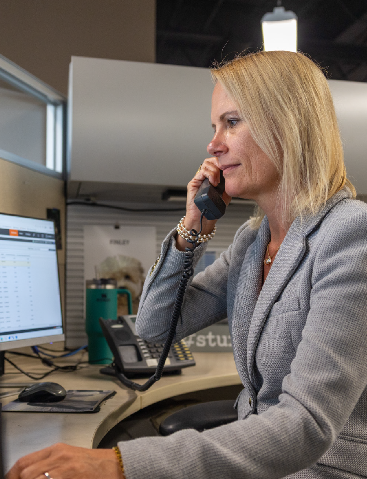 Woman in a gray blazer talking on a corded office phone while looking at a computer screen at her desk.