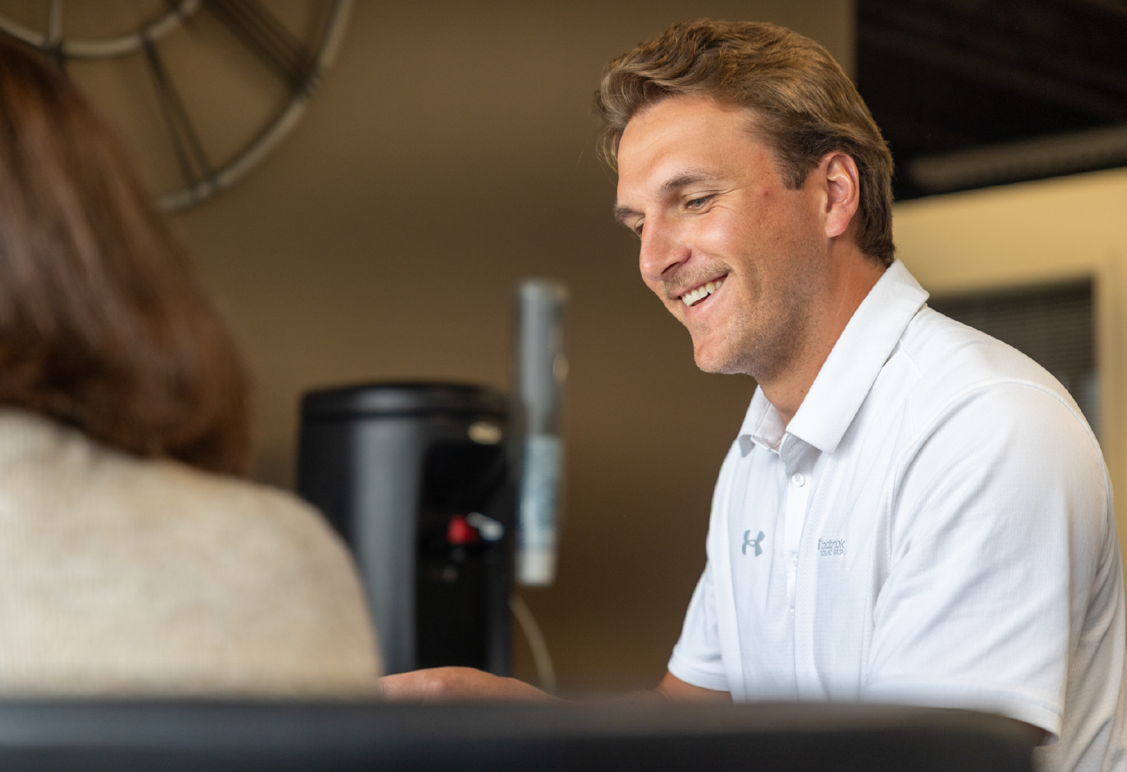 Smiling man wearing a white polo shirt sitting indoors, engaged in conversation with a person whose back is to the camera.