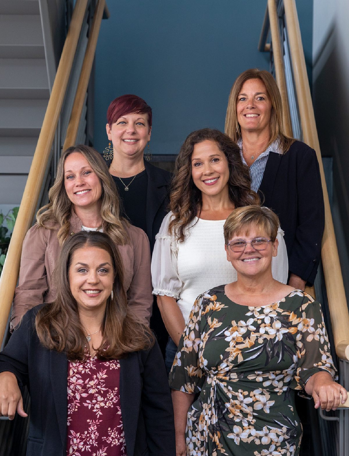 Six smiling women standing on a staircase with wooden railings in an indoor setting, wearing casual to business-casual attire.