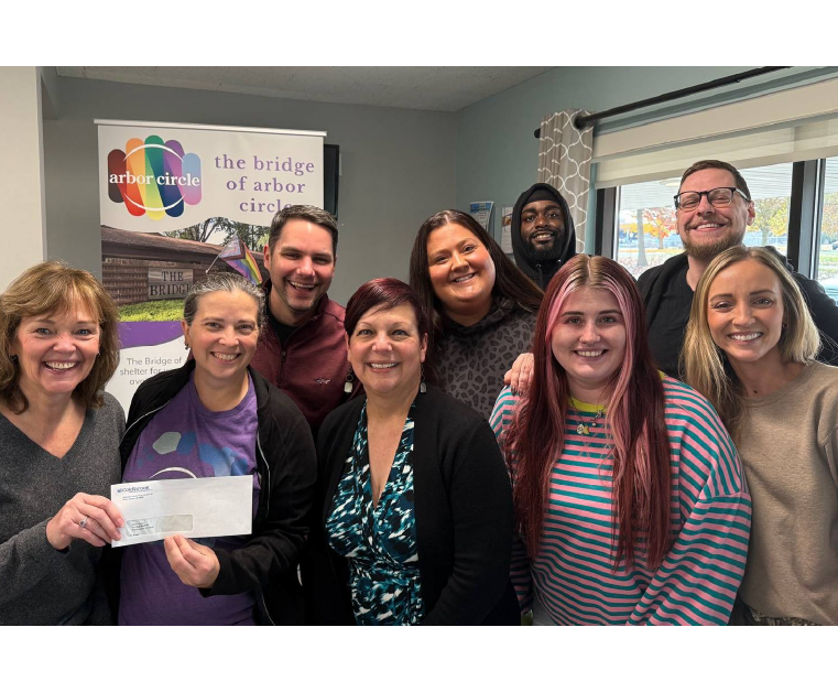 Group of nine diverse adults smiling indoors, standing close together, with one woman holding an envelope; a banner reading 'arbor circle' and 'the bridge of arbor circle' is visible in the background.