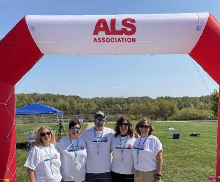 Five people wearing matching white ALS Association T-shirts standing together under a red and white inflatable arch with ALS Association written at the top, outdoors on green grass with a blue canopy and trees in the background.