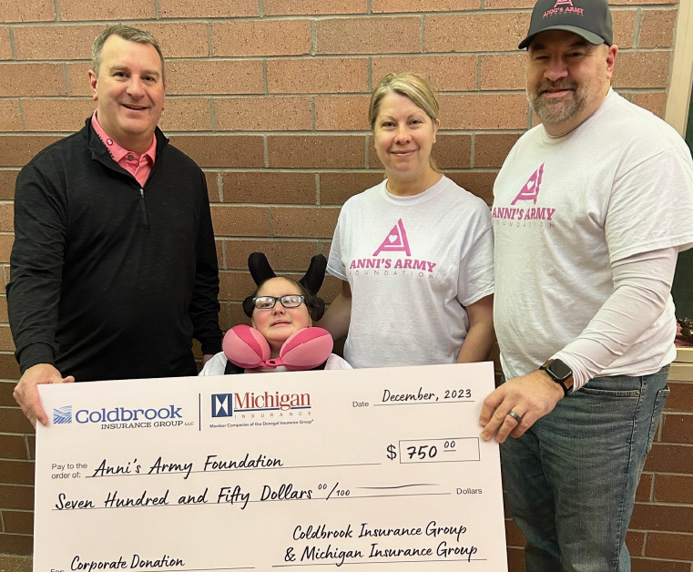 Four people standing in front of a brick wall holding a large check for $750 made out to Anni's Army Foundation from Coldbrook Insurance Group and Michigan Insurance Group as a corporate donation in December 2023. Two individuals wear white Anni's Army t-shirts, one person wears a black jacket, and a young person in a wheelchair with glasses and a pink neck pillow is in the center.