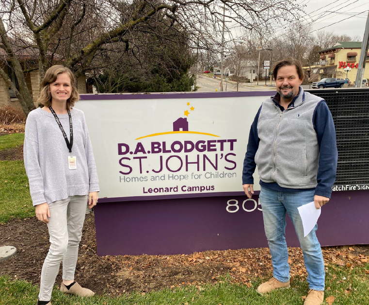 A woman and a man stand on either side of a large sign for D.A. Blodgett St. John's Leonard Campus, which provides homes and hope for children. The sign is purple and white with yellow stars above a house icon. The background shows trees and a street.