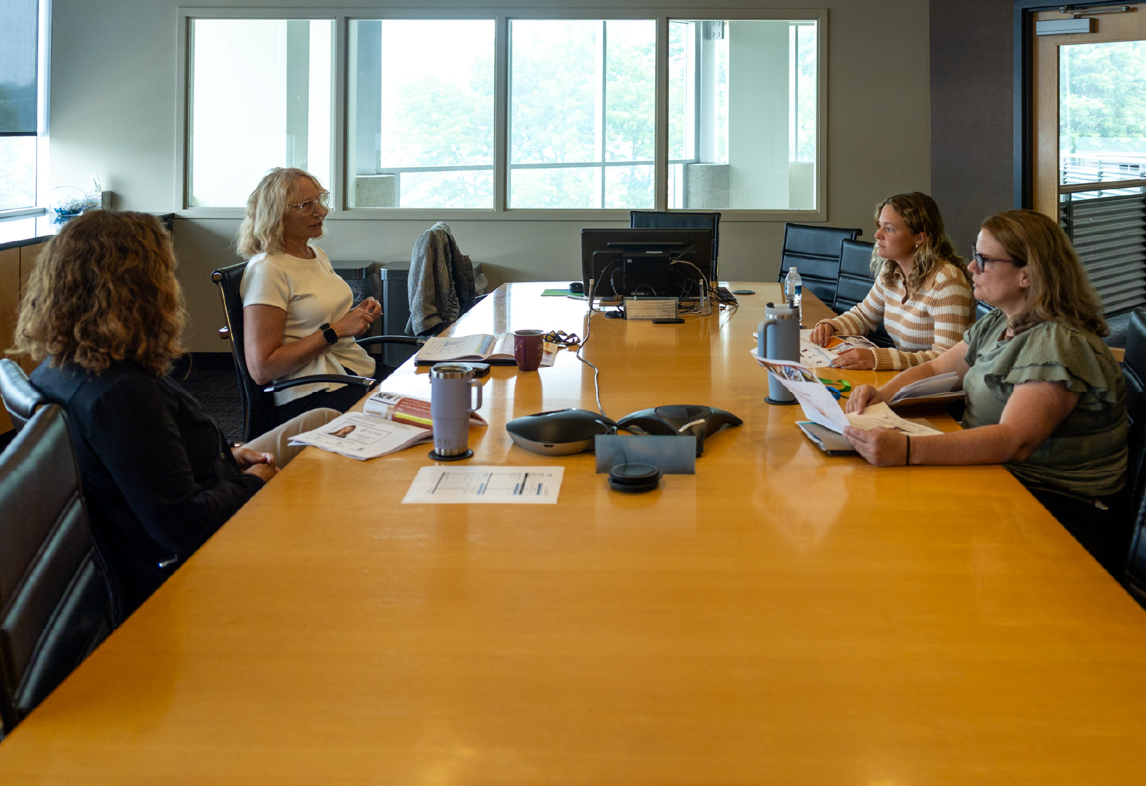 Four women seated around a large conference table in an office meeting room, engaged in discussion with papers, notebooks, and water bottles on the table.