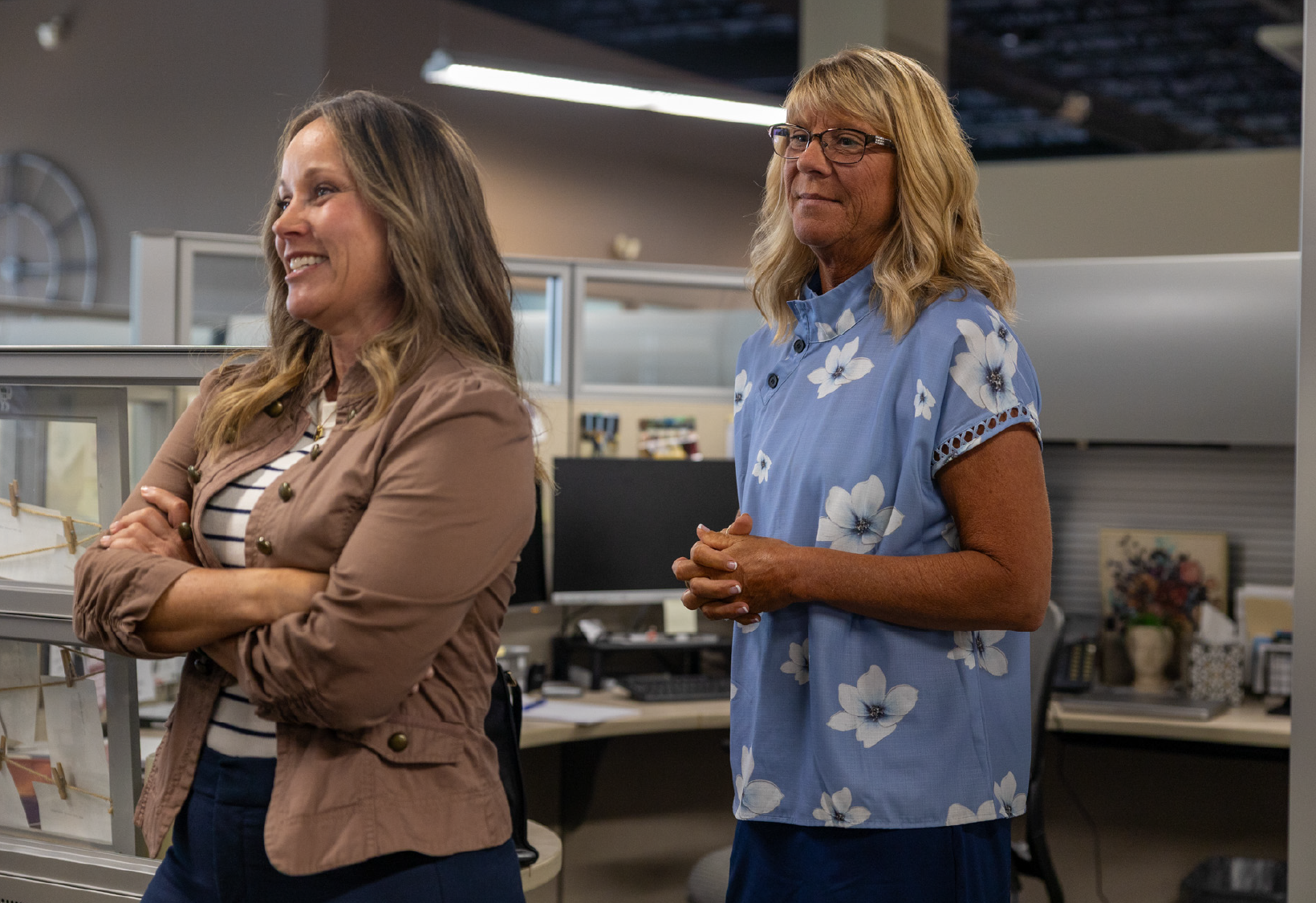 Two women standing in an office cubicle; the woman on the left smiles with crossed arms wearing a brown jacket over a striped shirt, and the woman on the right looks on wearing glasses and a blue floral blouse.
