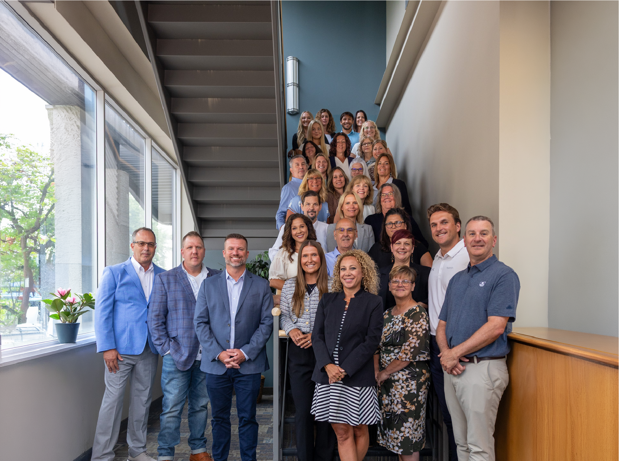 Group portrait of over 30 people standing and sitting on an indoor staircase, dressed in business casual attire, with large windows and a potted plant visible on the left side.