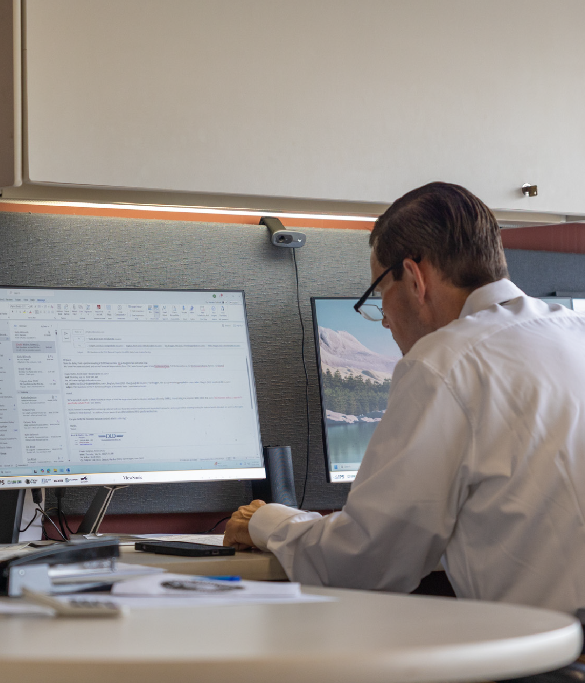 Man in a white shirt and glasses working at a desk with two monitors, one displaying an email inbox, in an office cubicle.