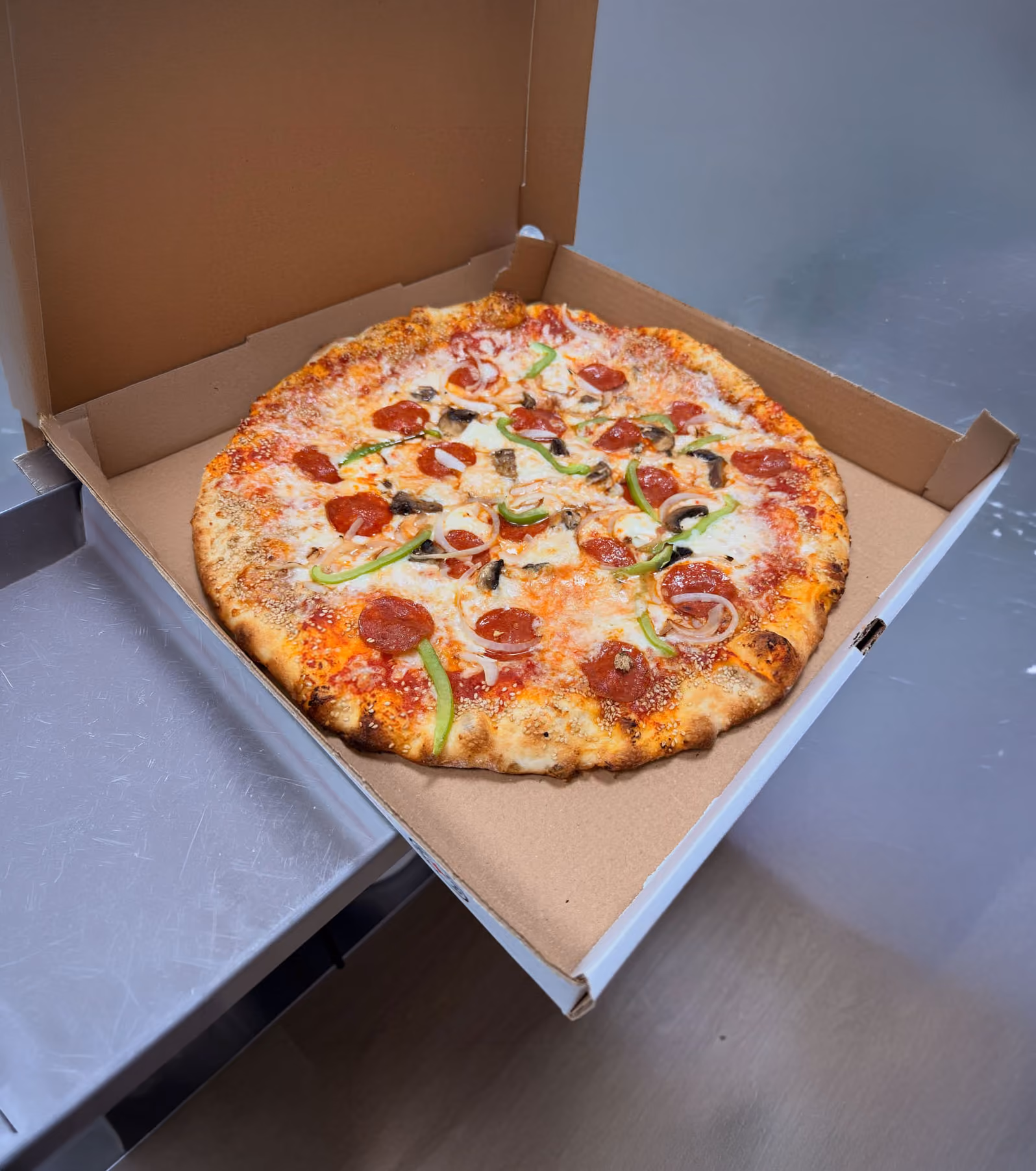 Pepperoni pizza with green peppers, mushrooms, and onions in an open cardboard box on a metal surface.