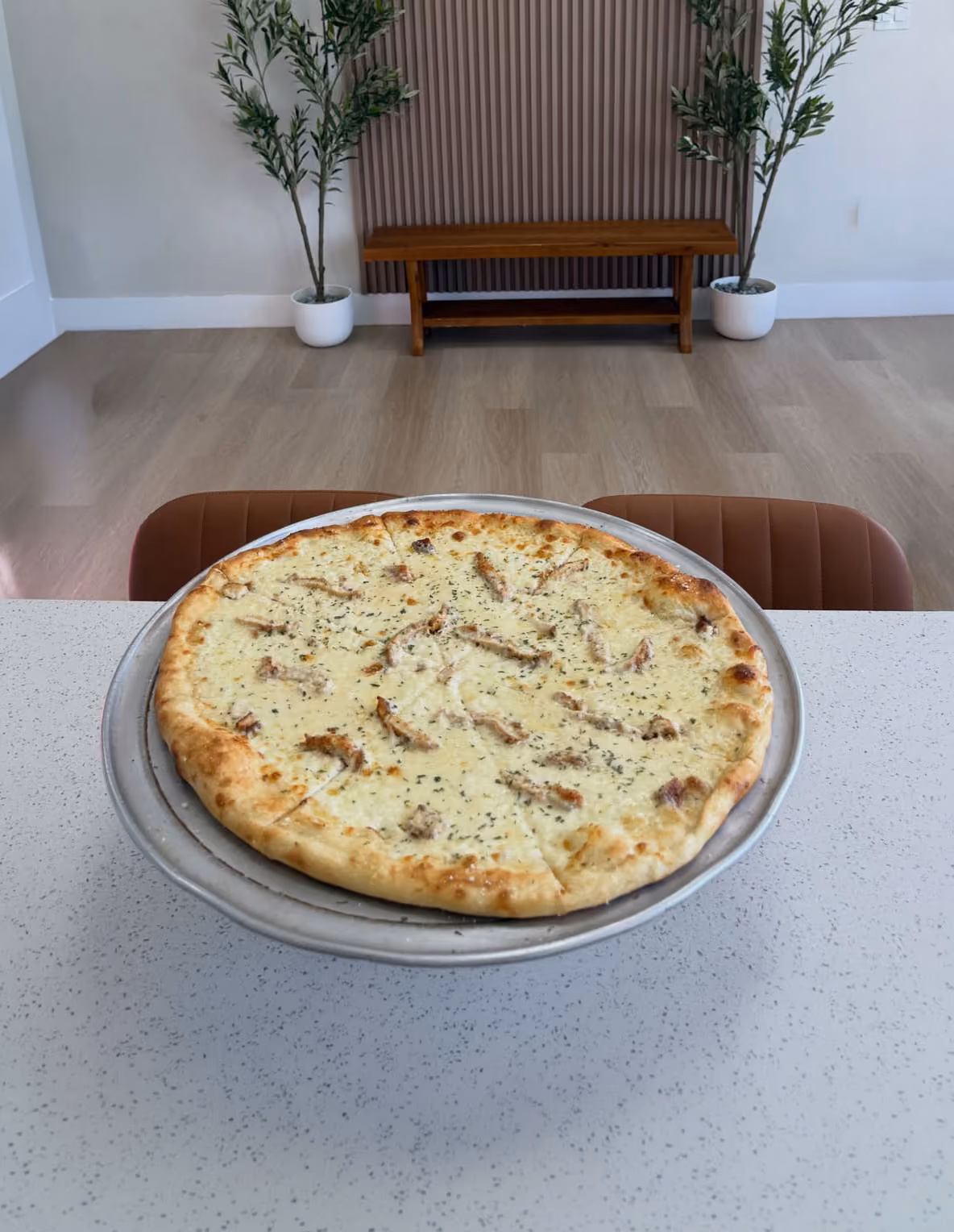 Whole white cheese pizza with chicken strips on a metal tray on a white speckled countertop.